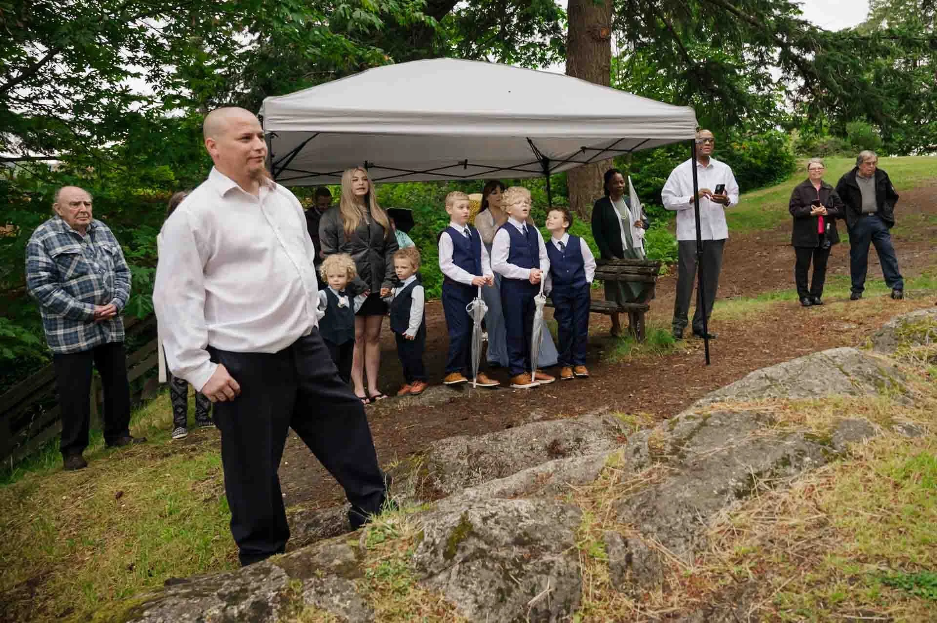 a group of people standing under a white tent in an open grassy area with trees behind them 