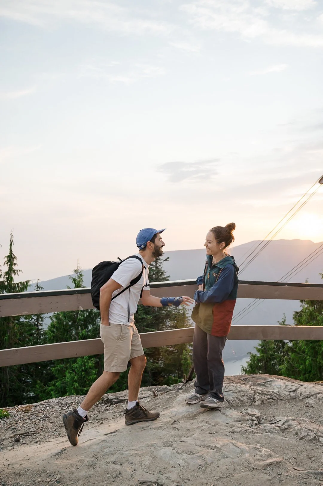 man and woman standing in front of a fence with trees and mountains in the background. man is bending forward and reaching into his pocket