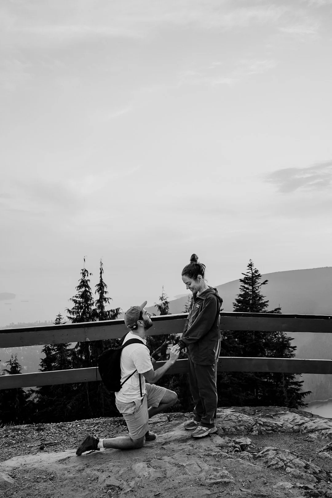 man proposing to woman in front of a wooden fence, with trees and a mountain in the background