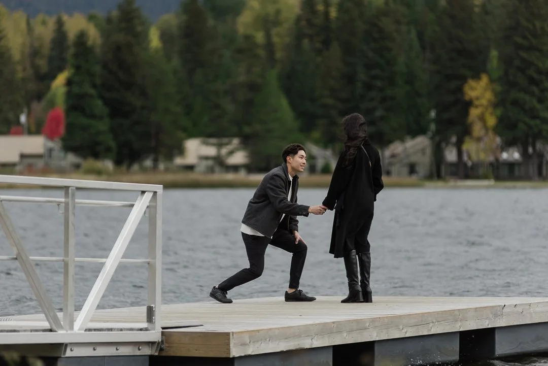 man and woman standing on a dock with water + trees in the background. man is bending down to get on one knee