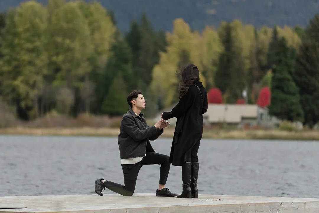 man down on one knee looking up at woman with water and trees in the background