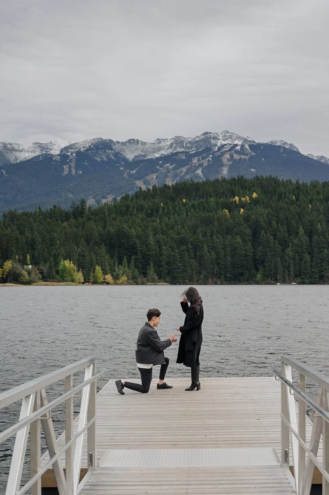 man proposing to woman at the end of a dock with water and mountains in the background