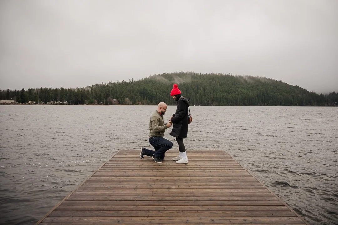 man on one knee in front of woman in black coat and red toque at the eod of a dock with water and mountains in the background