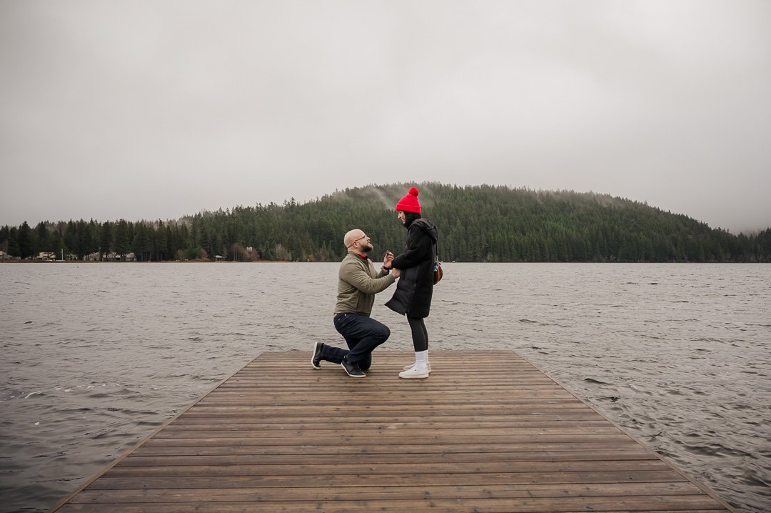 man holding a ring down on one knee looking up at woman with water and trees in the background