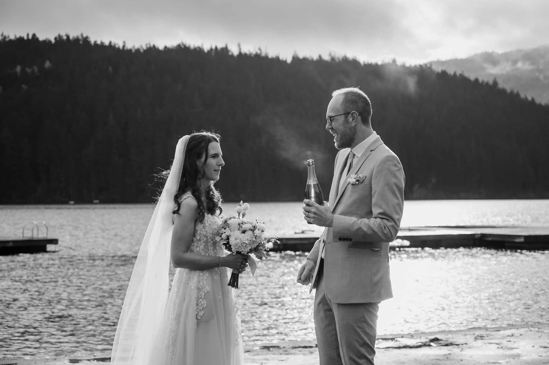 bride and groom opening a bottle of champagne by a lake with trees in the background