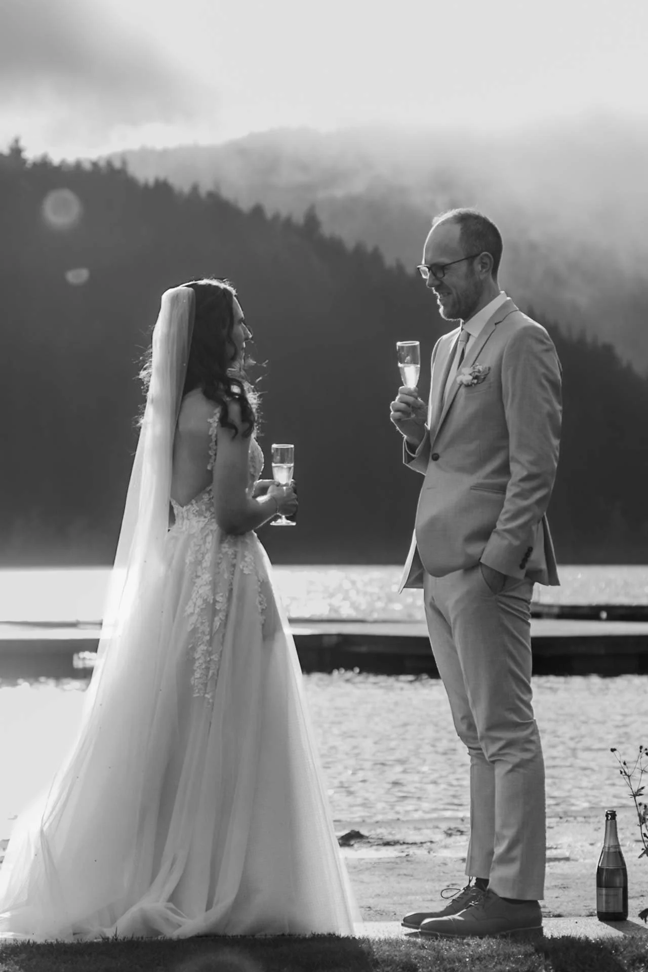 Couple enjoying a glass of champagne by the lake with mountains + trees in the background