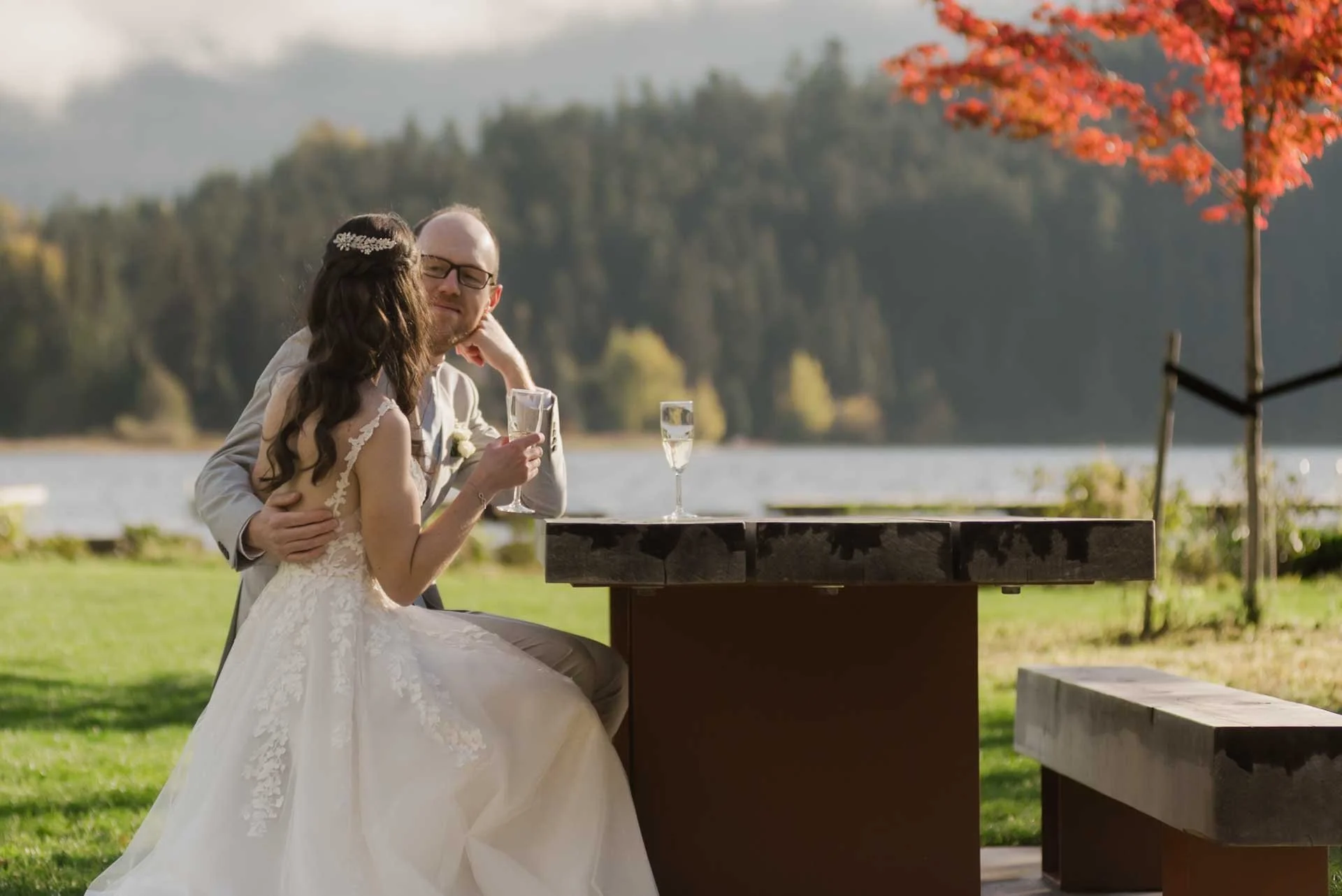 bride and groom sitting at a picnic table by the lake enjoying some bubbly
