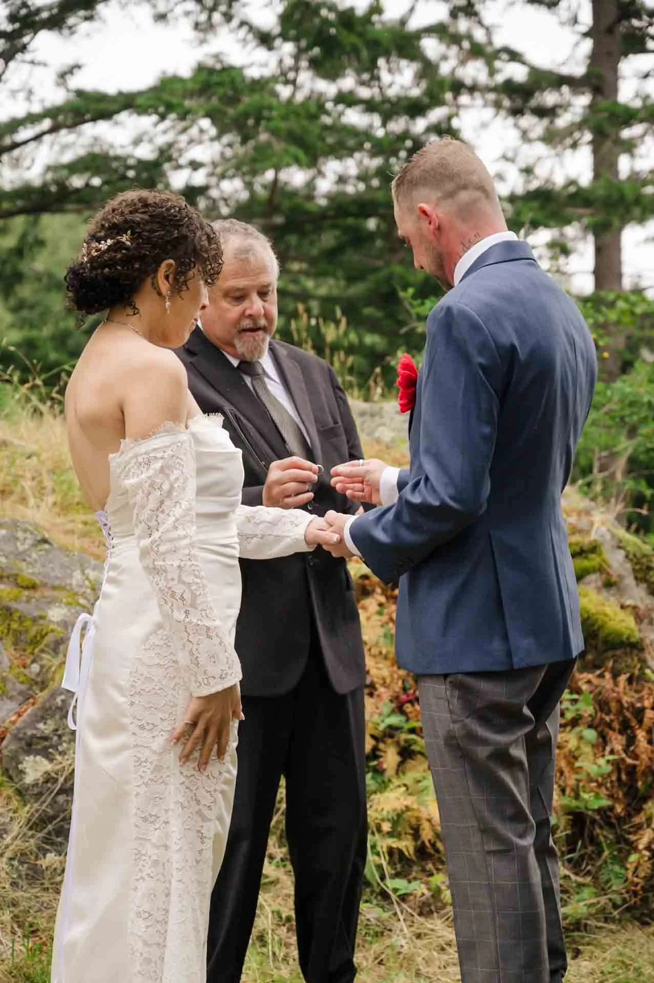 groom putting ring on bride's finger standing in front of an officiant in a park