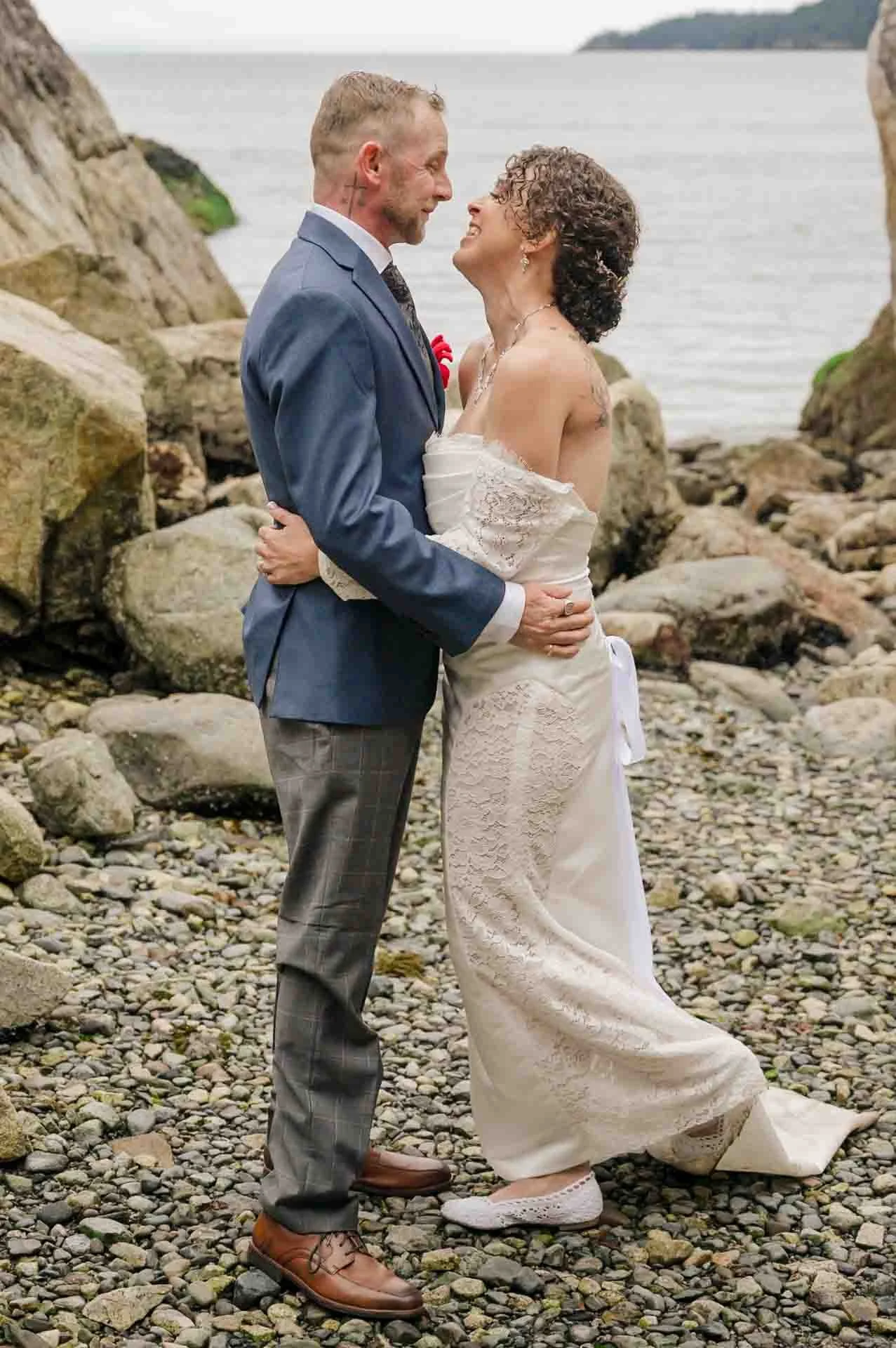 bride and groom facing each other, hugging, on a rocky beach in front of the ocean