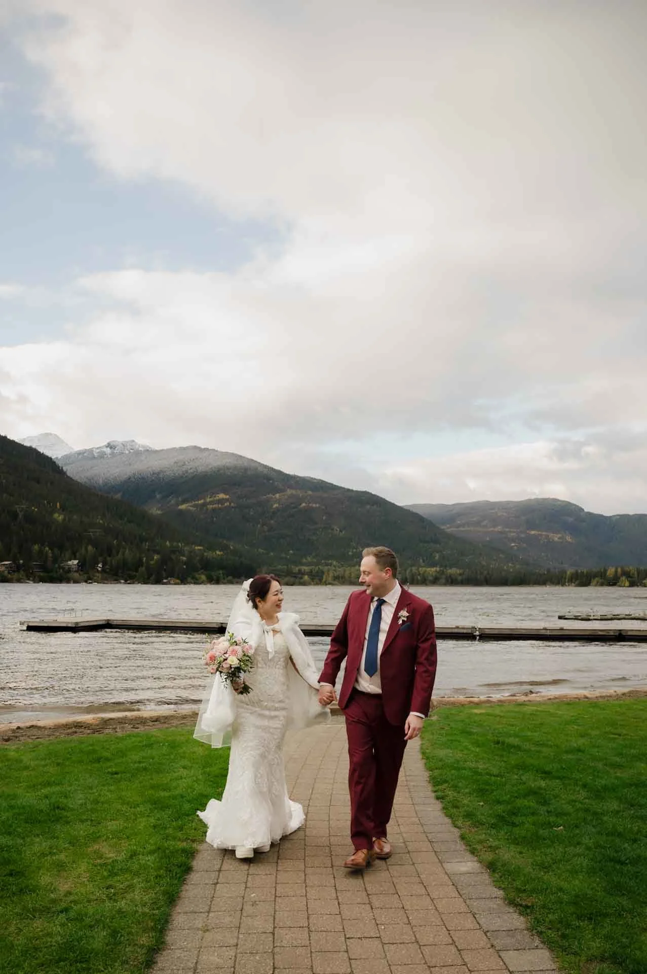 bride and groom holding hands walking on a path in front of a lake and mountains