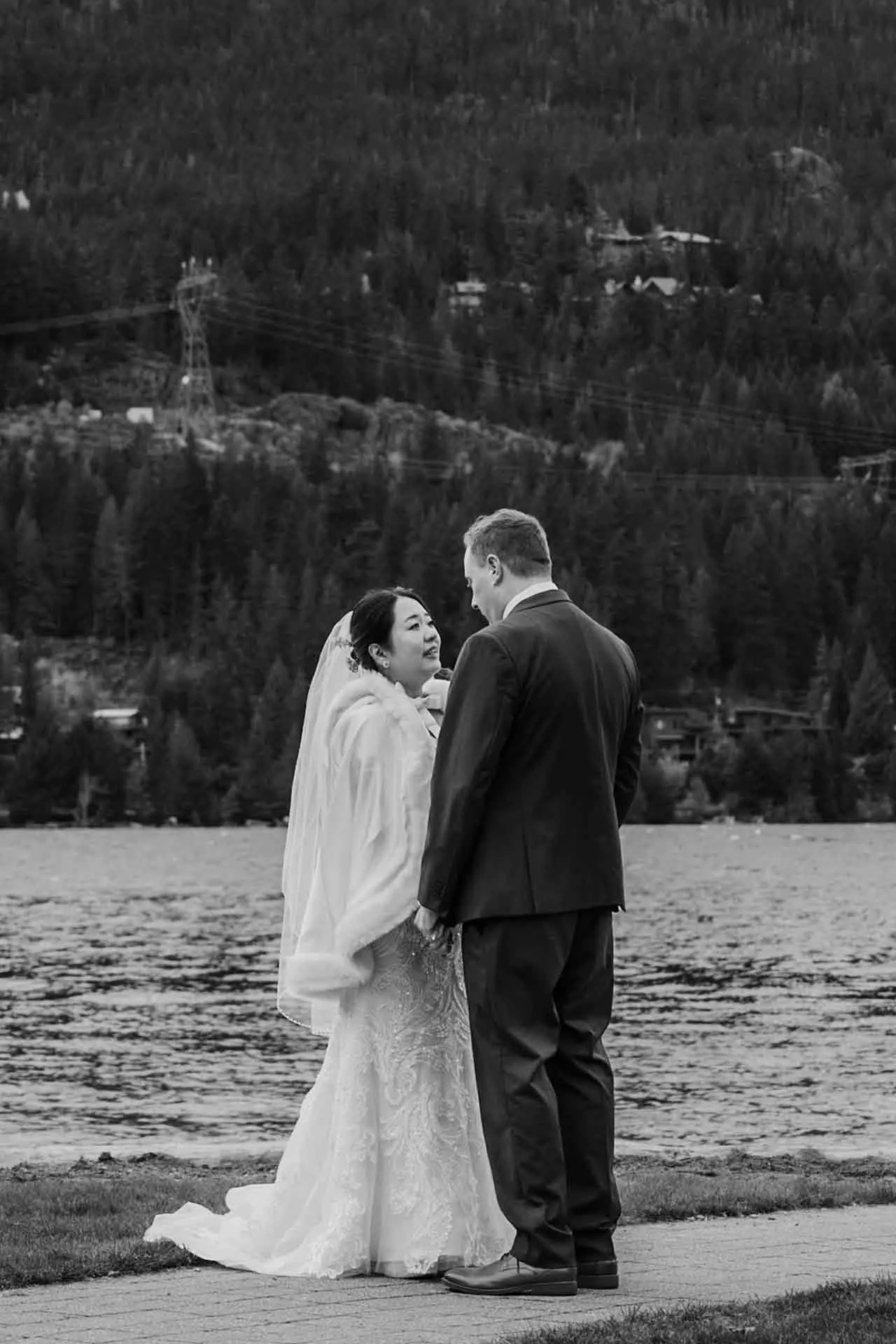 bride + groom facing eachother holding hads in front of a lake + mountains in the background