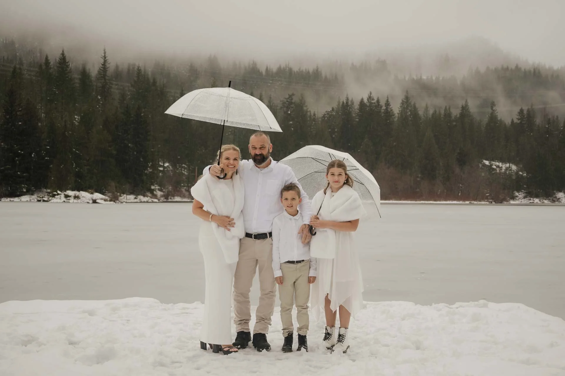 Bride + groom with their kids standing on a snowy dock with a frozen lake, green fir trees with low clouds, holding clear umbrellas
