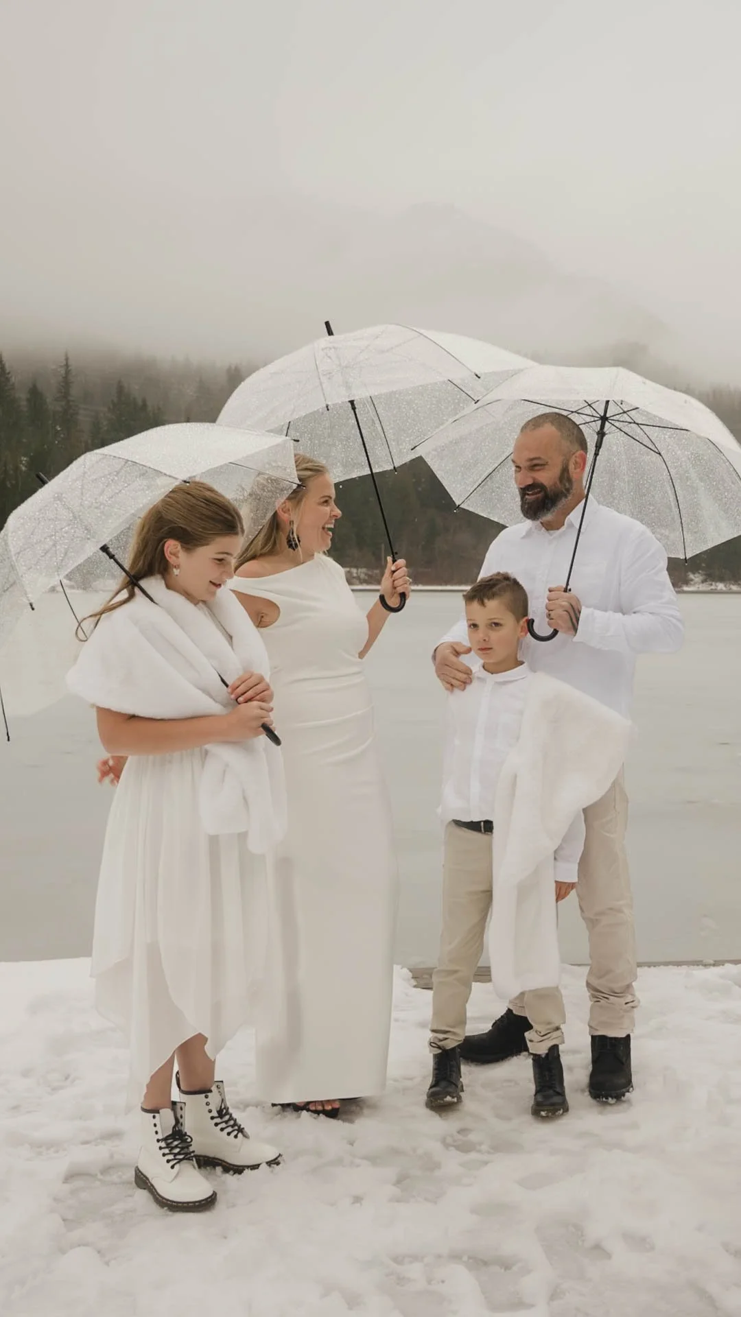 A couple eloping with their kids all standing on a snow-covered dock on a frozen lake holding umbrellas