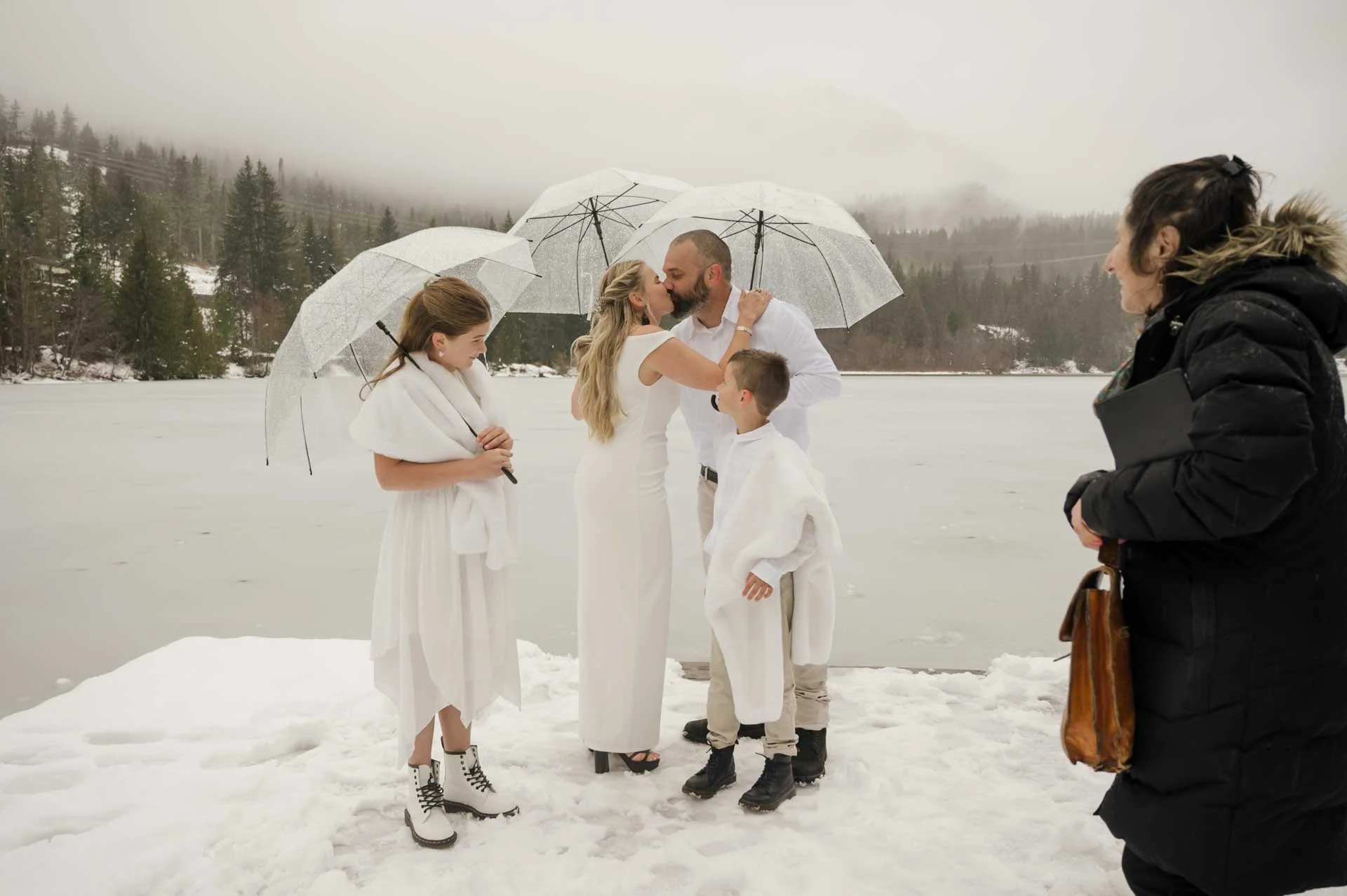 bride and groom kissing under clear umbrellas while their kids + officiant looking. everyone is standing on a snowy dock on a frozen lake with mountains + low clouds in the background