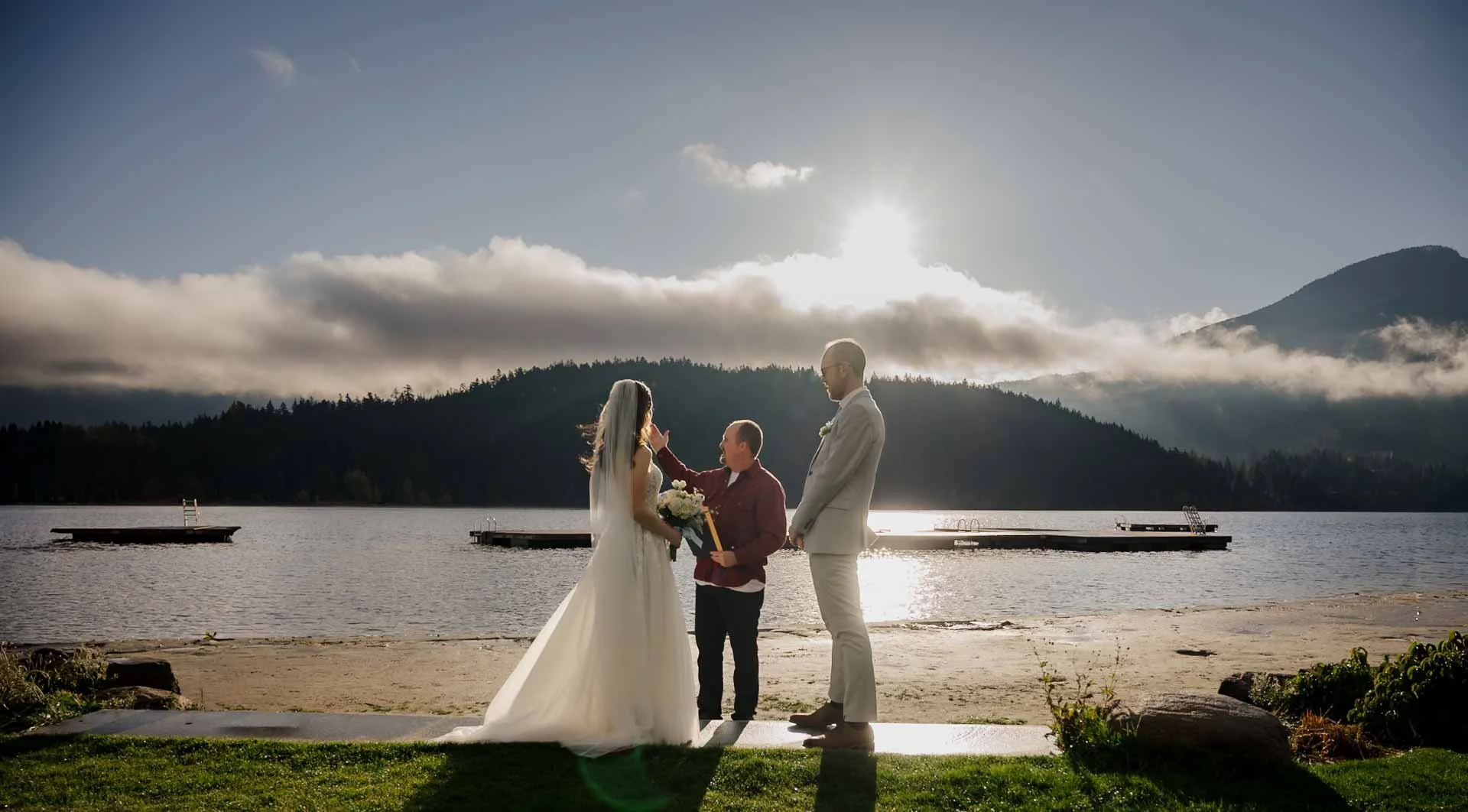 bride and groom and officiant standing on a beach in front of a lake with mountains in the background.