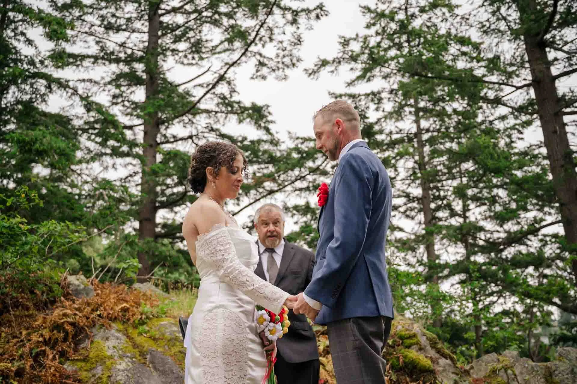 man and woman holding hands with a wedding officiant in front of them, and trees and rocks in the background