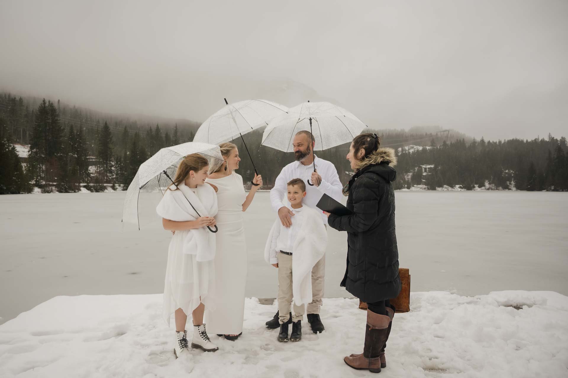 A couple dressed in white with their kids, also dressed in white, holding clear umbrellas on a dock with a frozen lake in the background and snow covered mountain,  with officiant