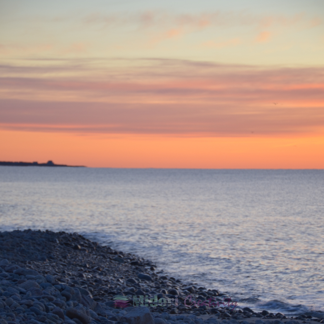 rocky shoreline beside calm ocean water under a soft pastel sunset sky with layered clouds