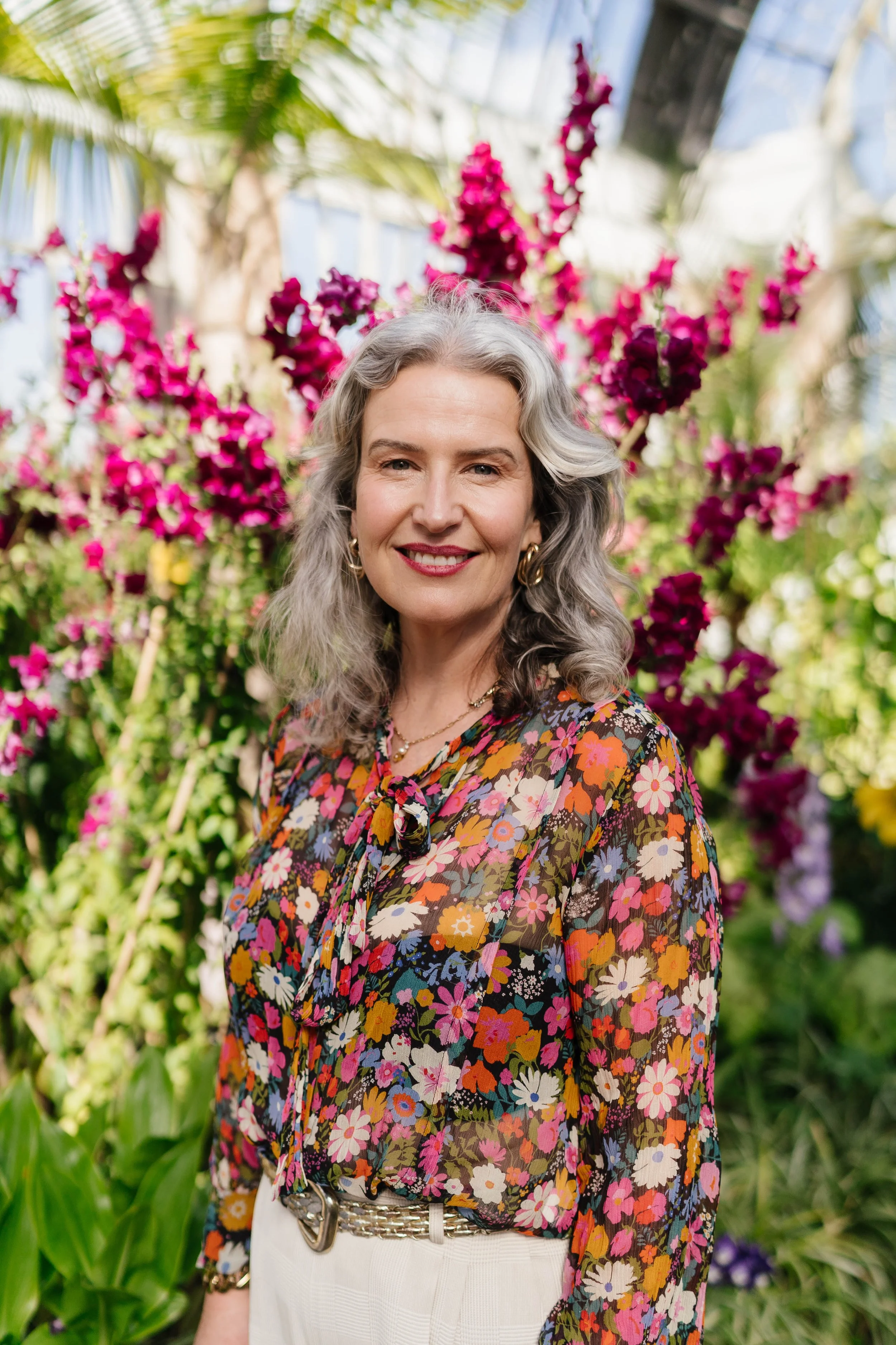 Older woman with gray, wavy hair smiling, wearing a colorful floral blouse, standing in a greenhouse with vibrant pink and purple flowers and green plants in the background.