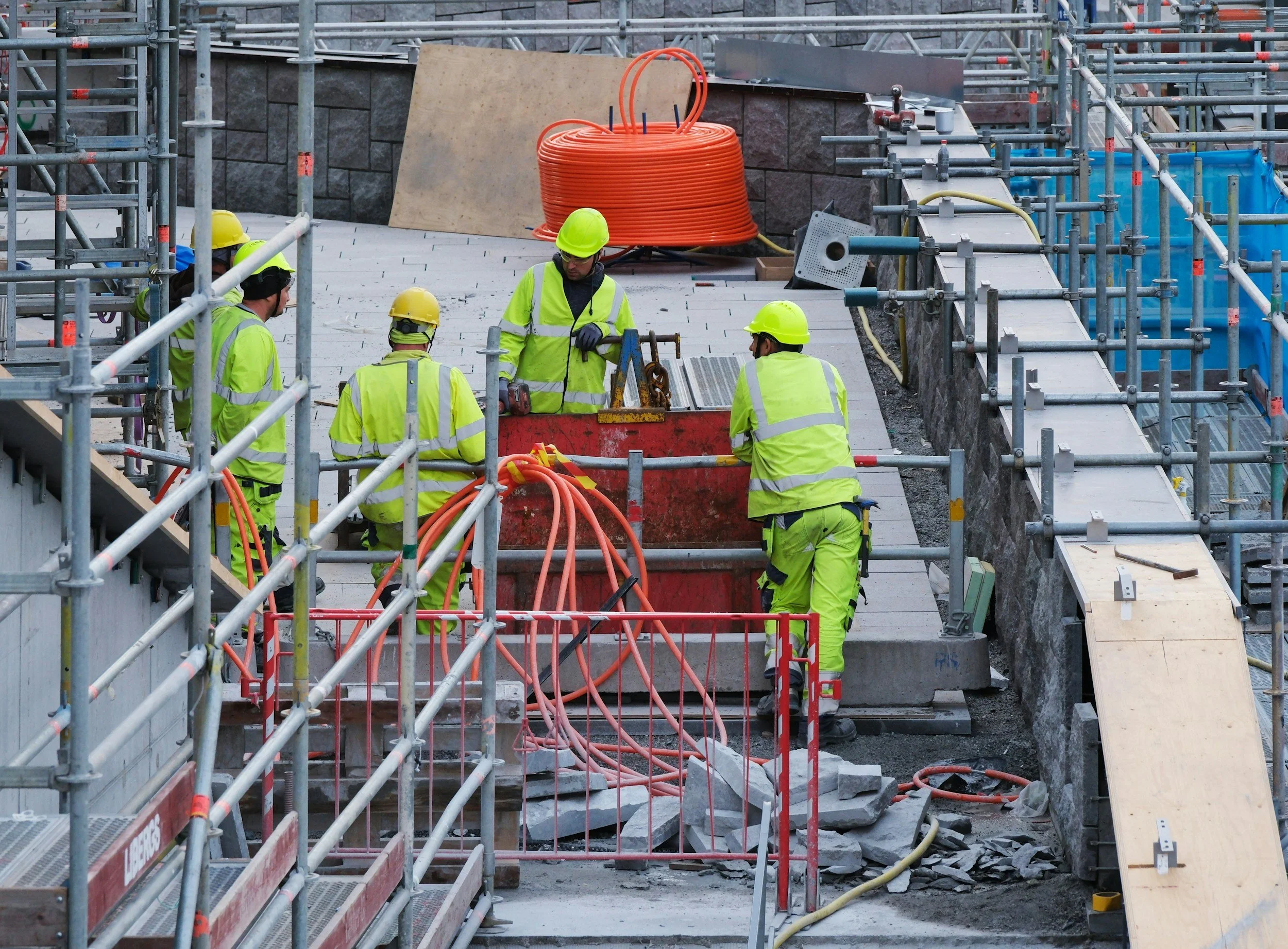 Construction workers wearing yellow safety helmets and neon yellow safety vests working on a construction site with scaffolding, orange hoses, and unfinished stone walls.
