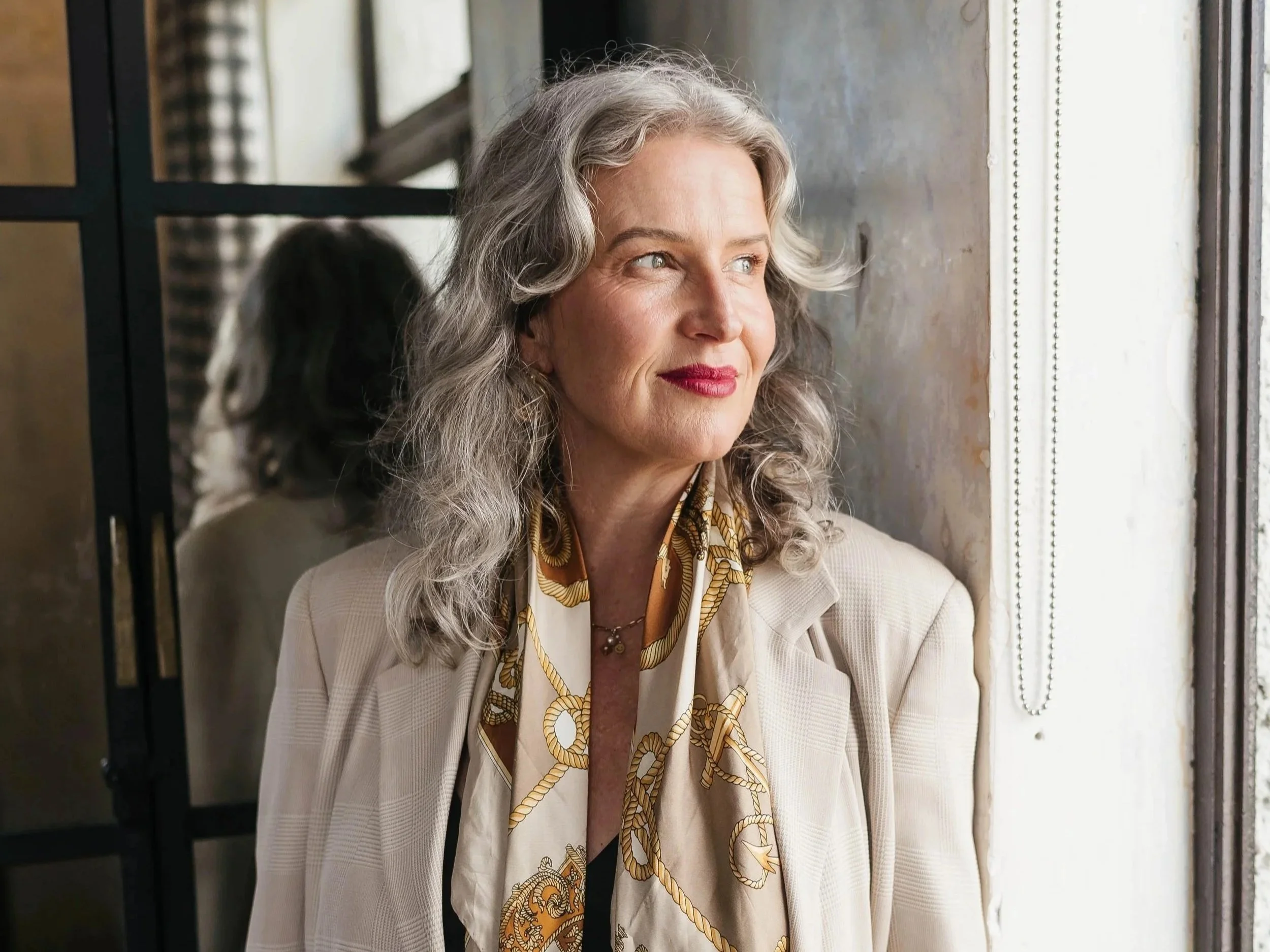 A mature woman with gray, curly hair looking out of a window with a thoughtful expression, wearing a beige blazer and a patterned silk scarf.