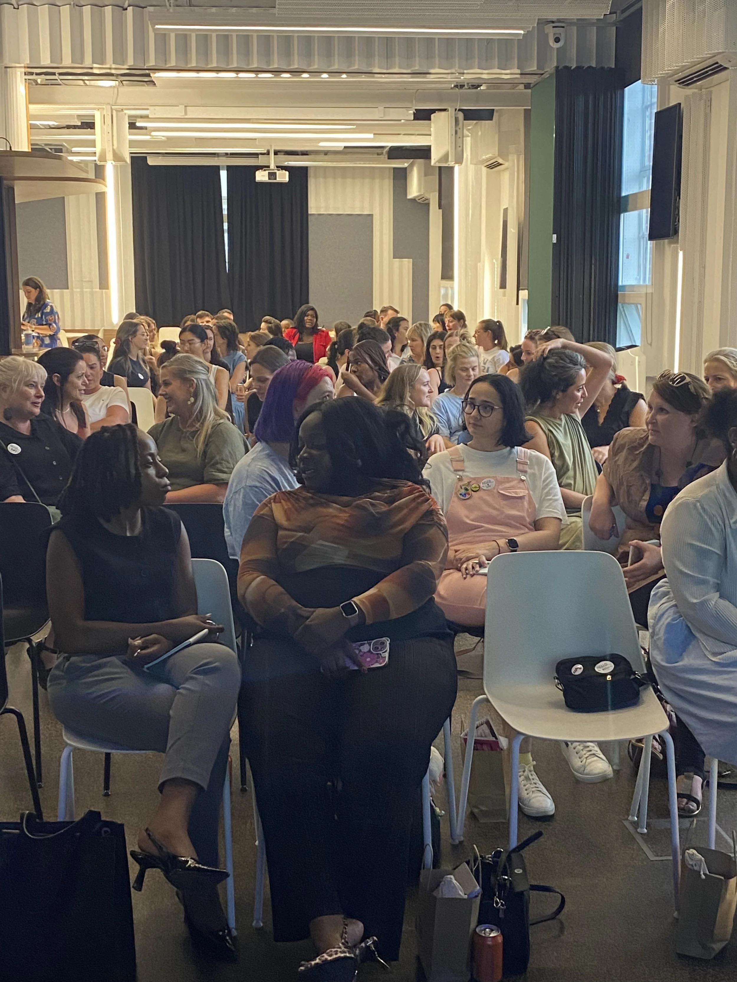 A diverse group of women attending a presentation or seminar inside a conference room with large windows, curtains, and a projector mounted on the ceiling.
