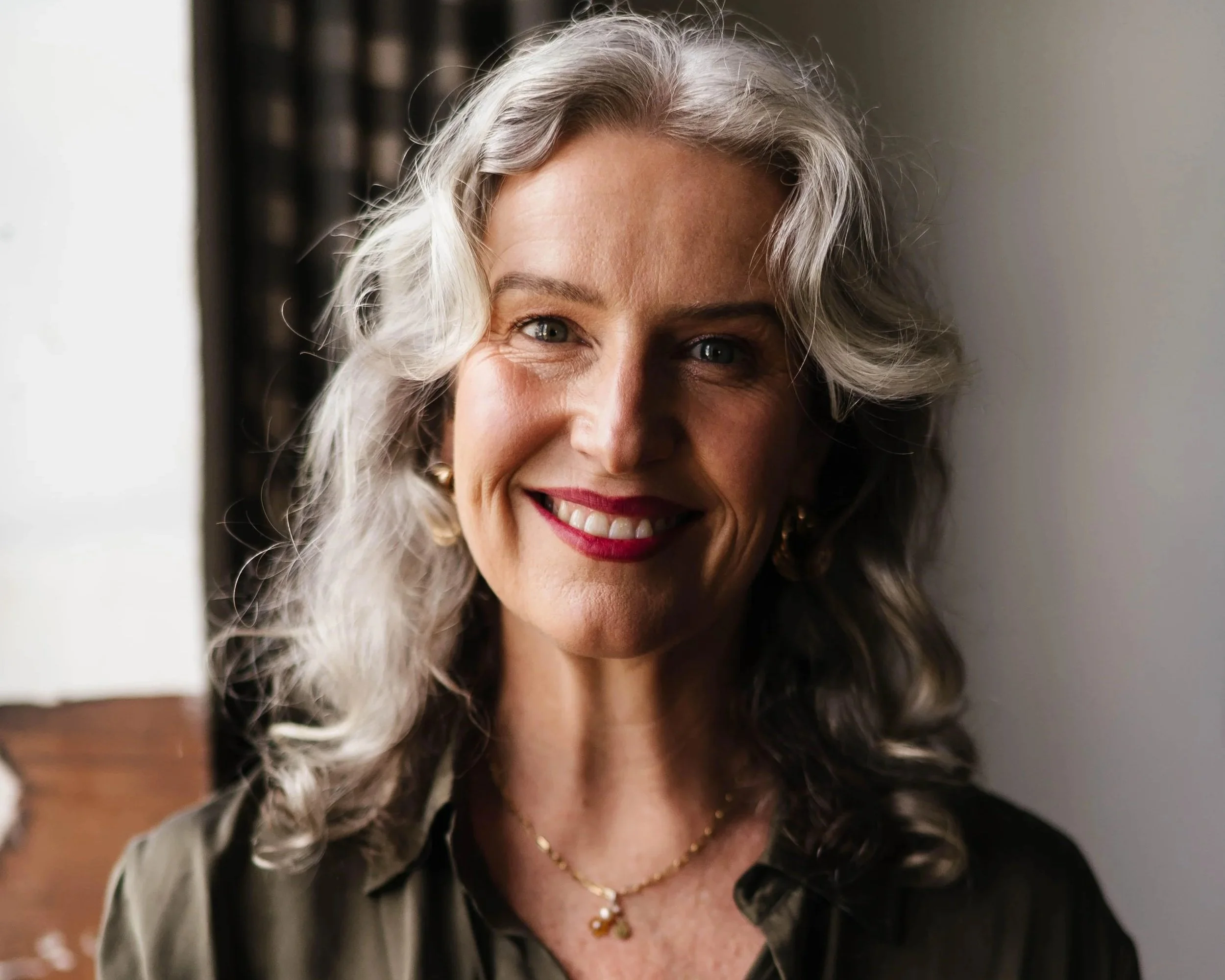 A smiling elderly woman with curly silver hair, wearing earrings and a gold necklace, in front of a plain background.