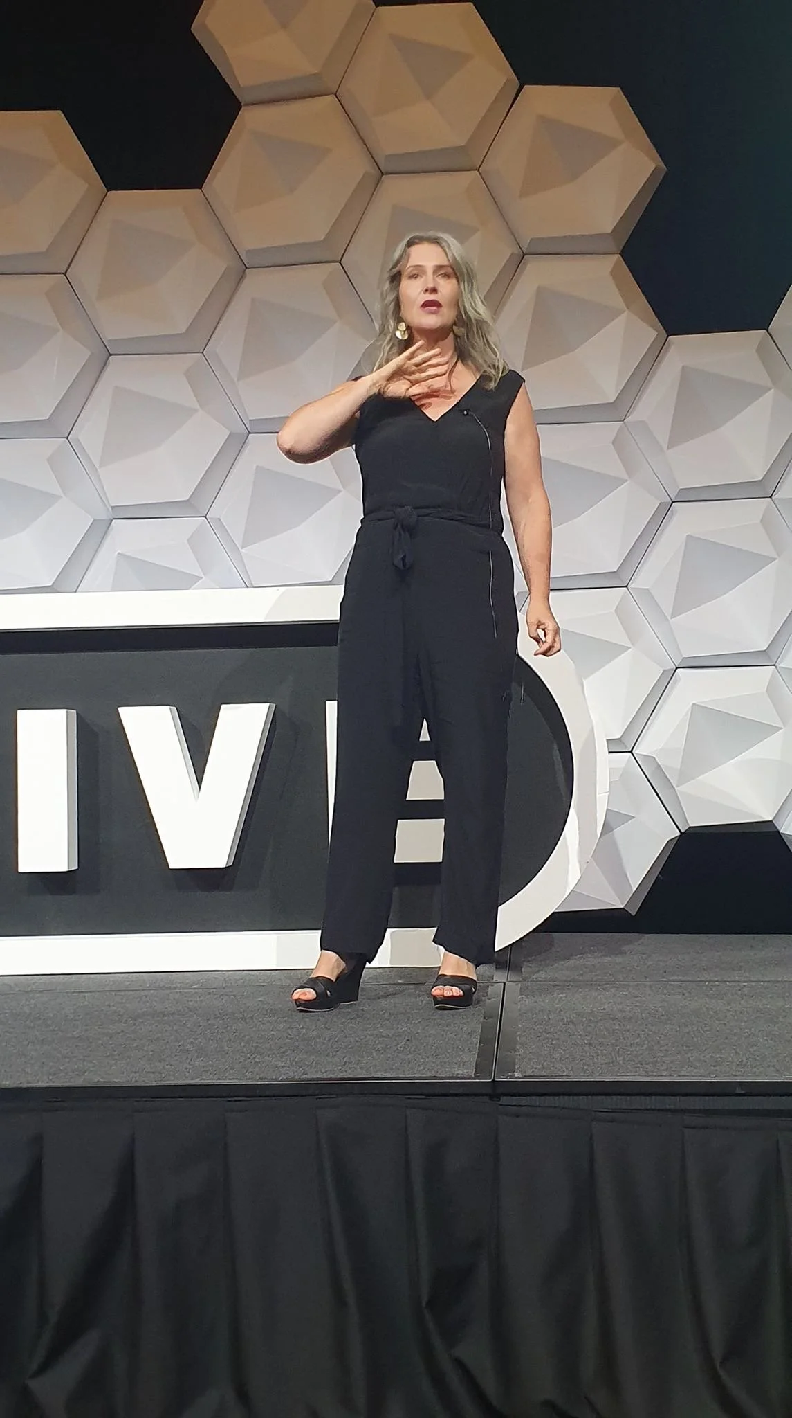 A woman with gray hair wearing a black sleeveless jumpsuit and open-toed black high heels standing on a stage with a black and white decorative background and large letters spelling WIB.
