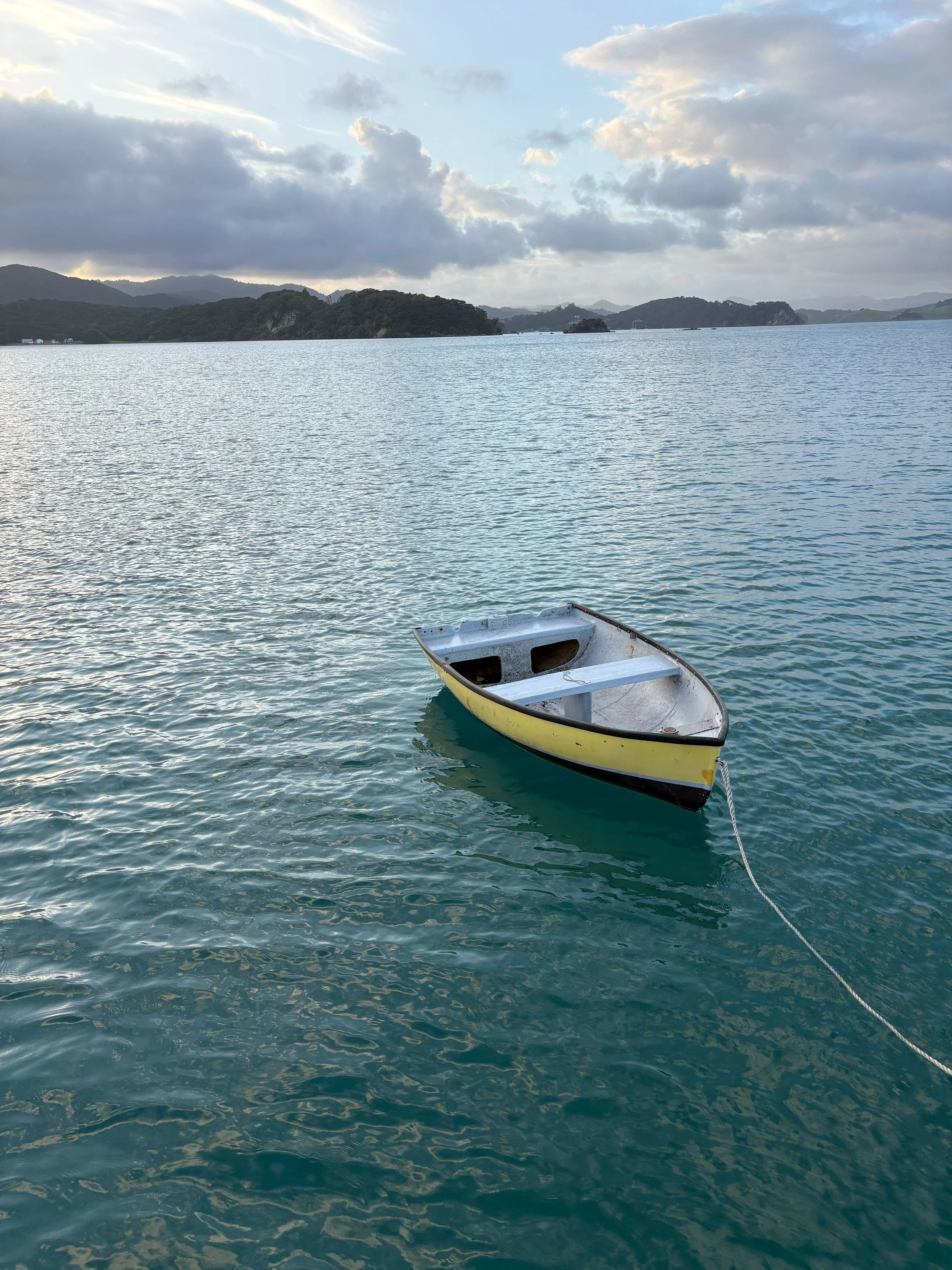 A lone yellow and white rowboat floating on calm blue water near a shoreline with hills and cloudy sky in the distance.