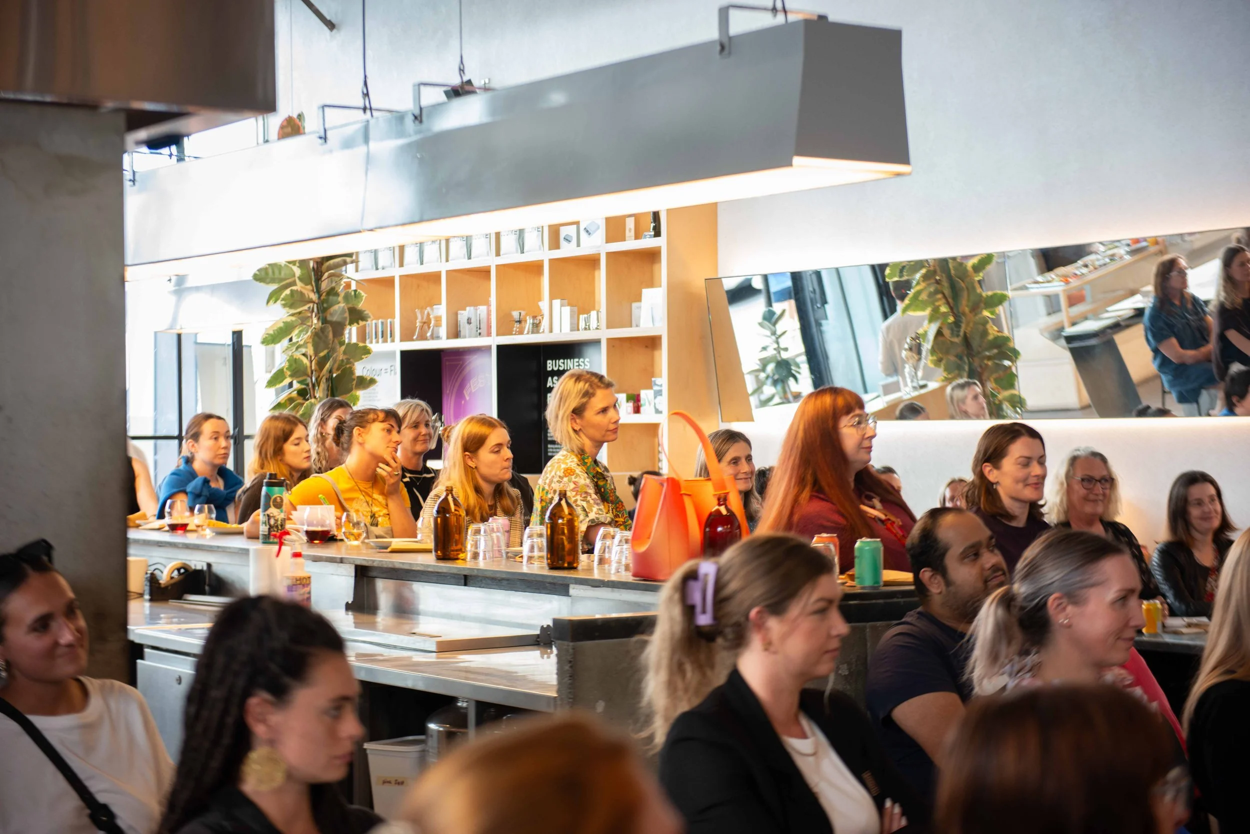 A group of people sitting at tables in an indoor event space, listening attentively. The setting features modern decor with large windows, plants, and shelves with books and signs. The image captures a diverse audience engaged in the event.