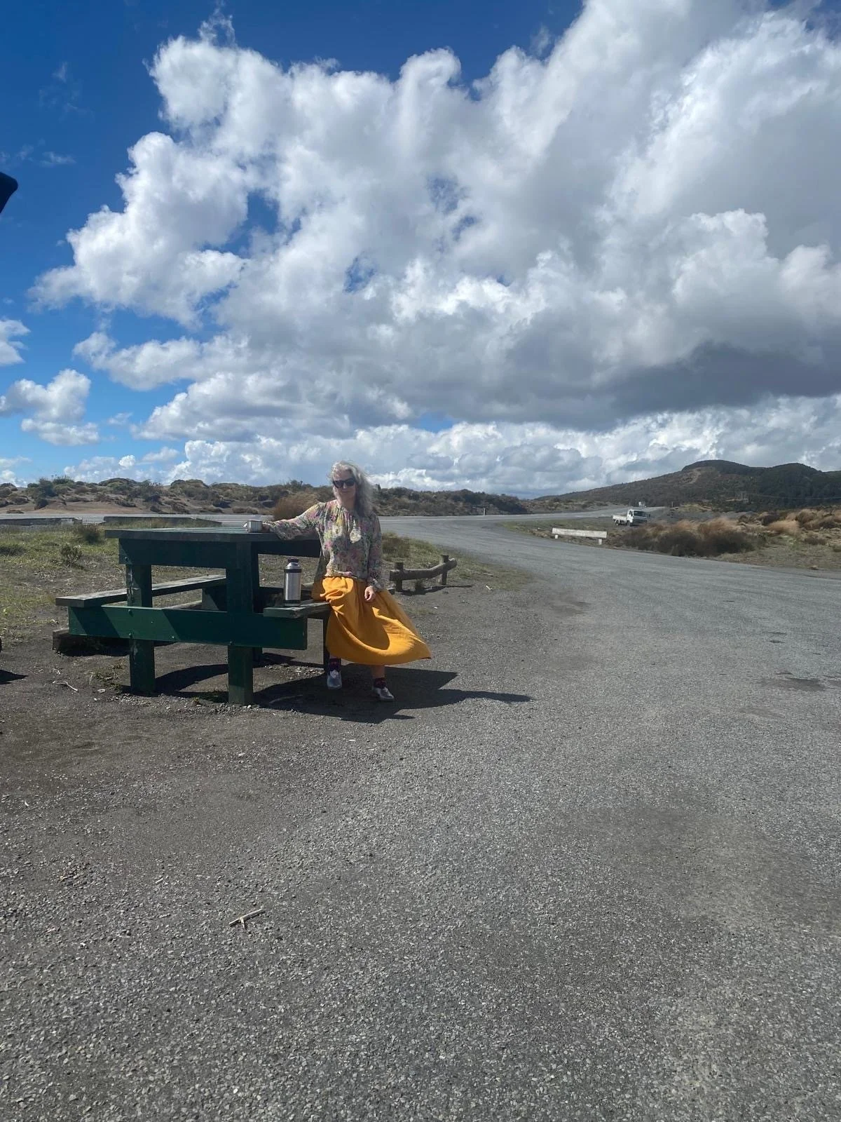 A woman in a floral shirt and long yellow skirt sitting at a green picnic table on a gravel area near a mountain road, with clouds and mountains in the background.