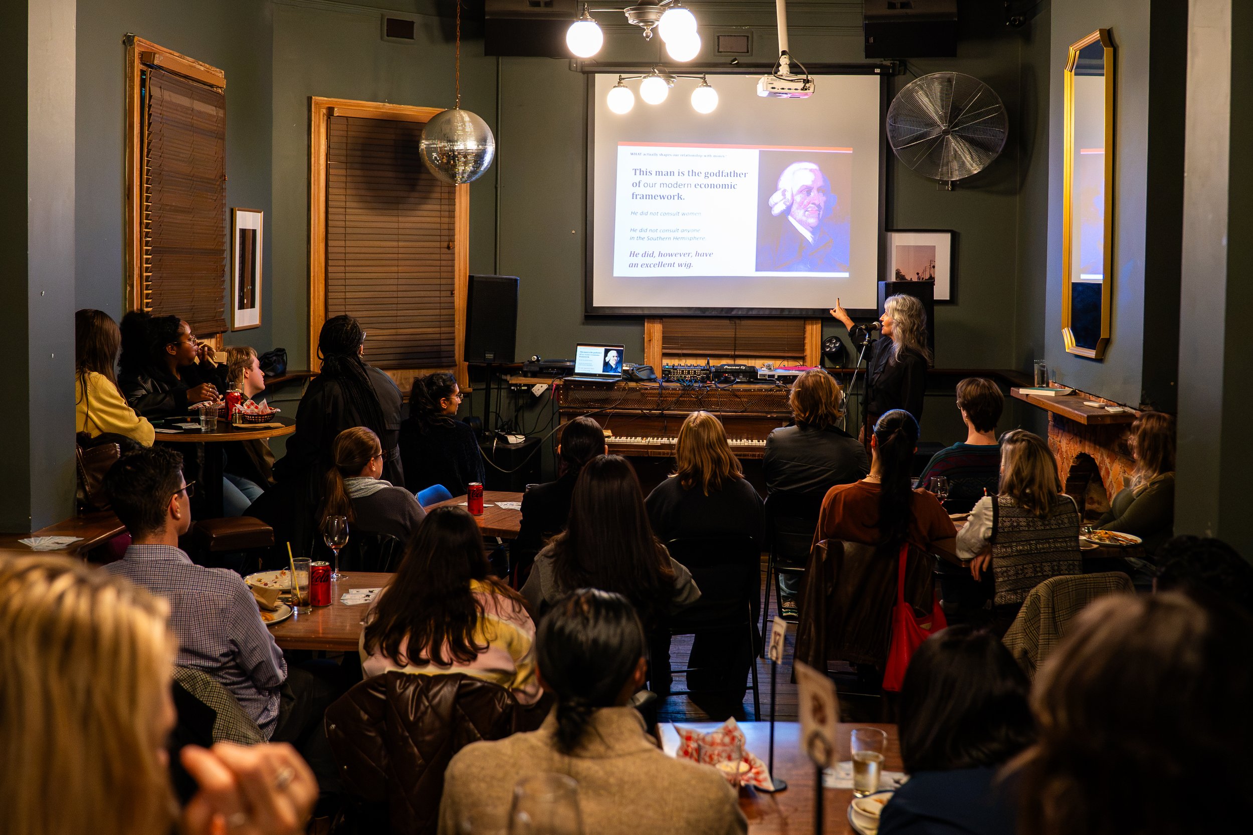 A group of people attending a presentation in a cozy, dimly lit room. A woman stands at the front, pointing to a projected slide on the screen, which features text and an image of Adam Smith. The audience is seated at tables, watching and listening.