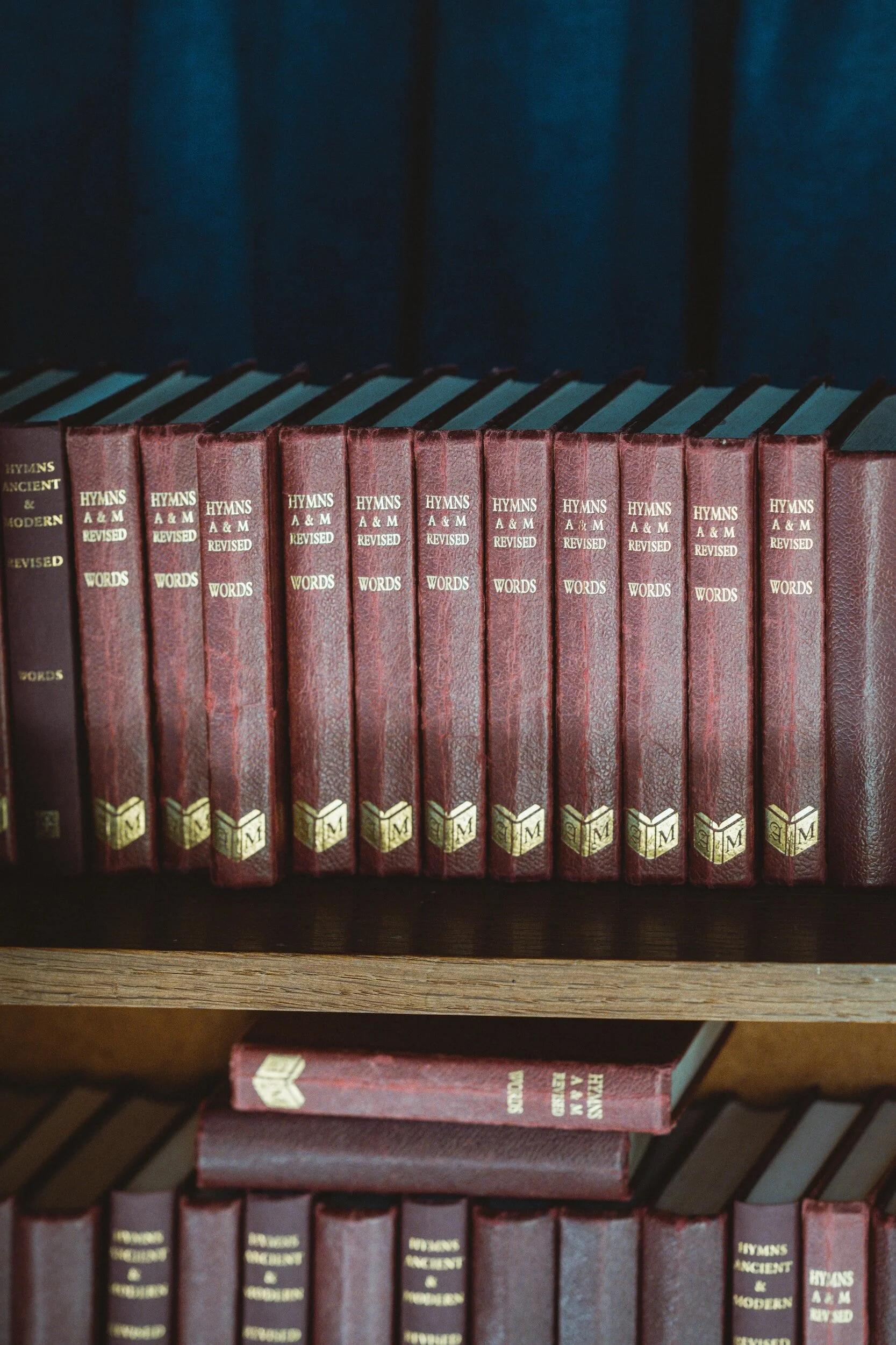 A row of hardcover books with red spines on a wooden shelf, titled 'Hymns A & M Revised Words'.