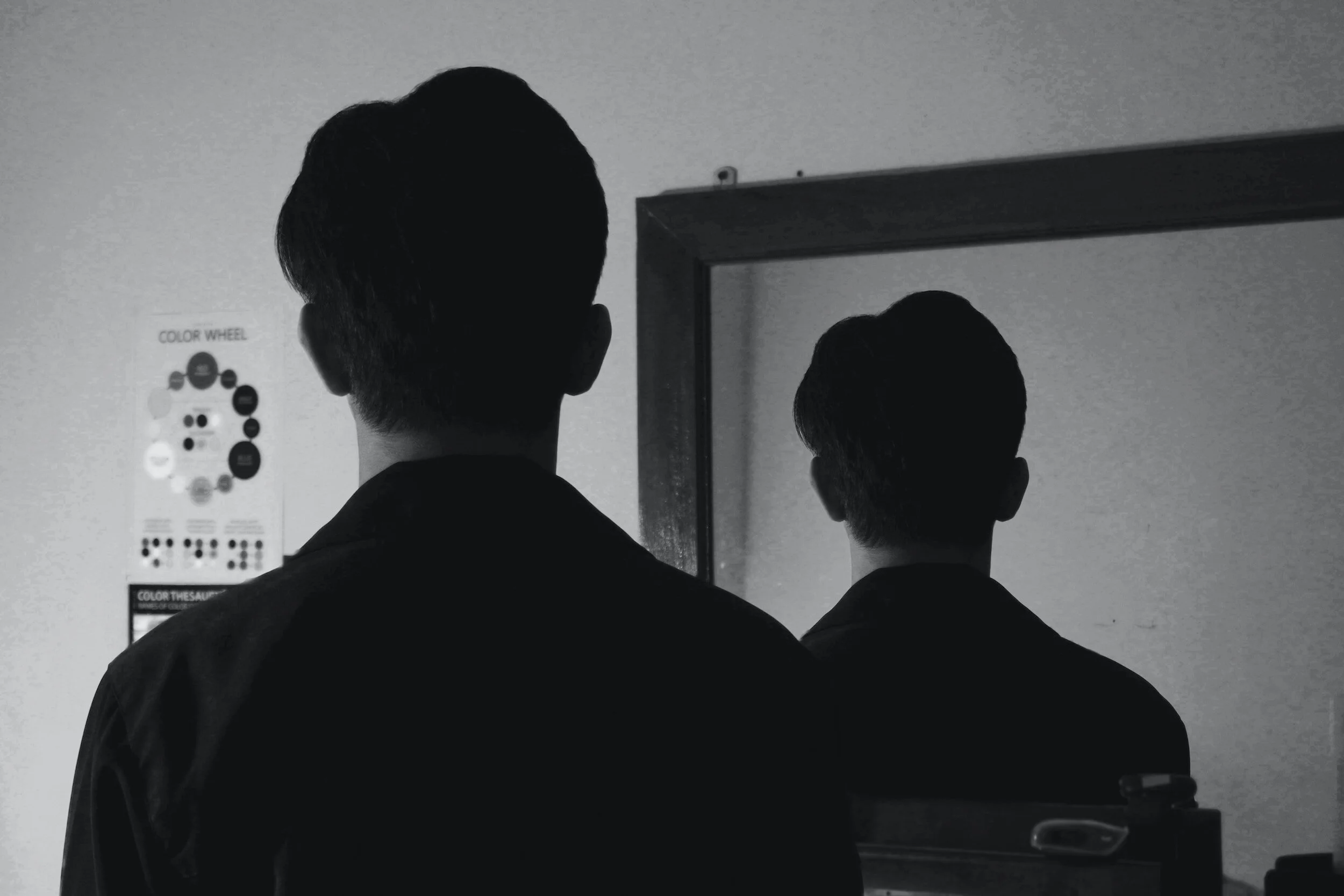Two individuals are looking at a mirror in a black-and-white photograph, with their backs facing the camera. The scene appears to be indoors, with a poster on the wall that includes color-related graphics.