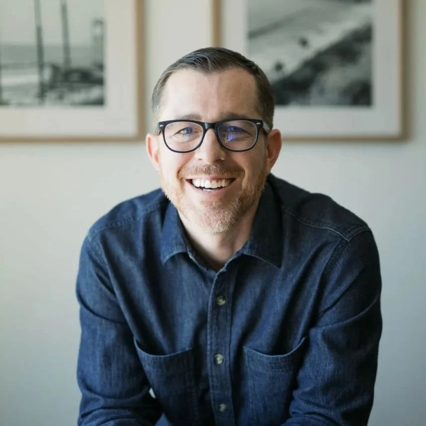 A man with short brown hair and blue eyes smiling, wearing a dark collared shirt, with a bookshelf and a colorful painting in the background.