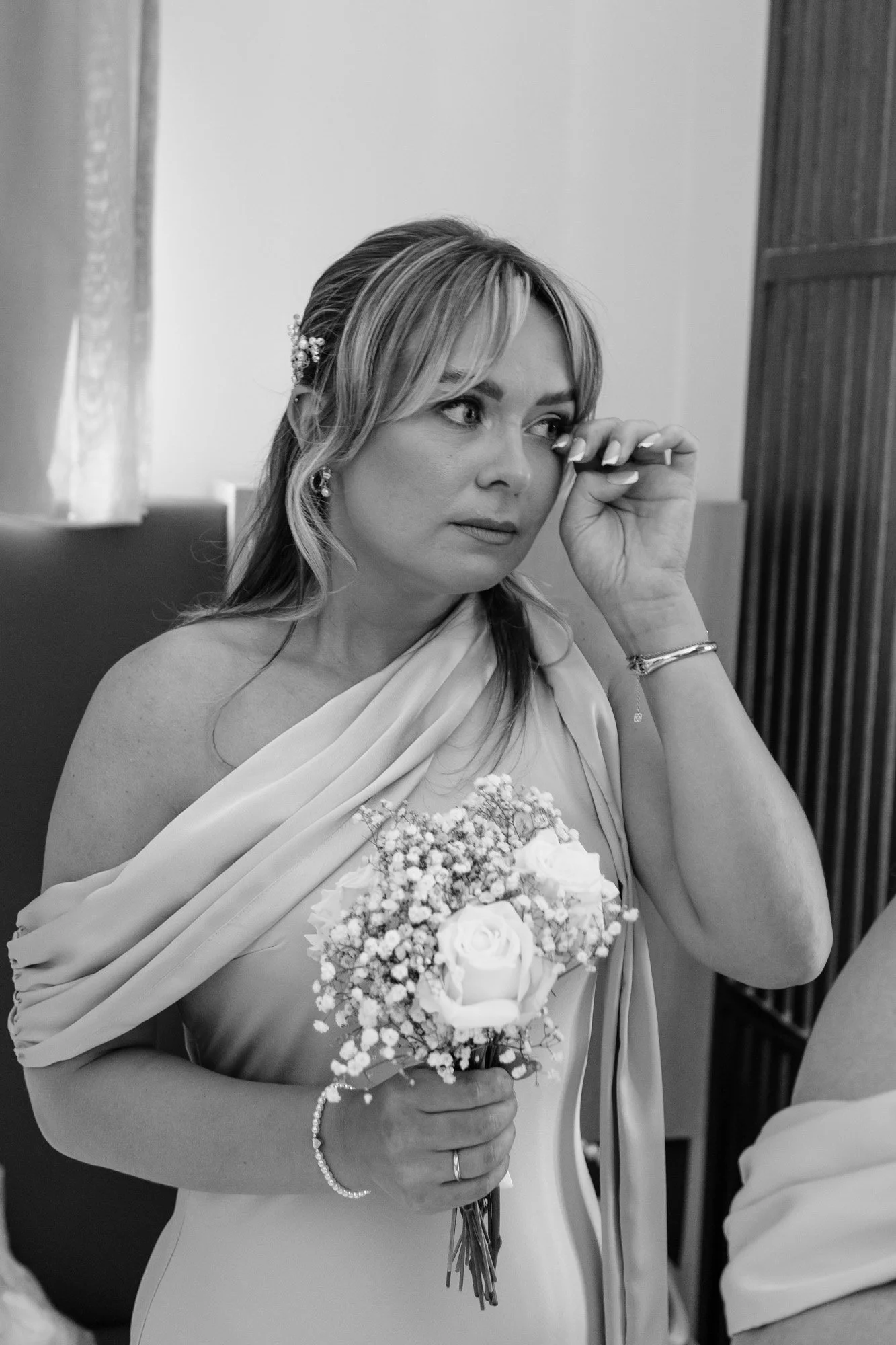 A woman dressed in wedding attire, holding a bouquet of flowers, appears to be emotional during a moment of the ceremony.