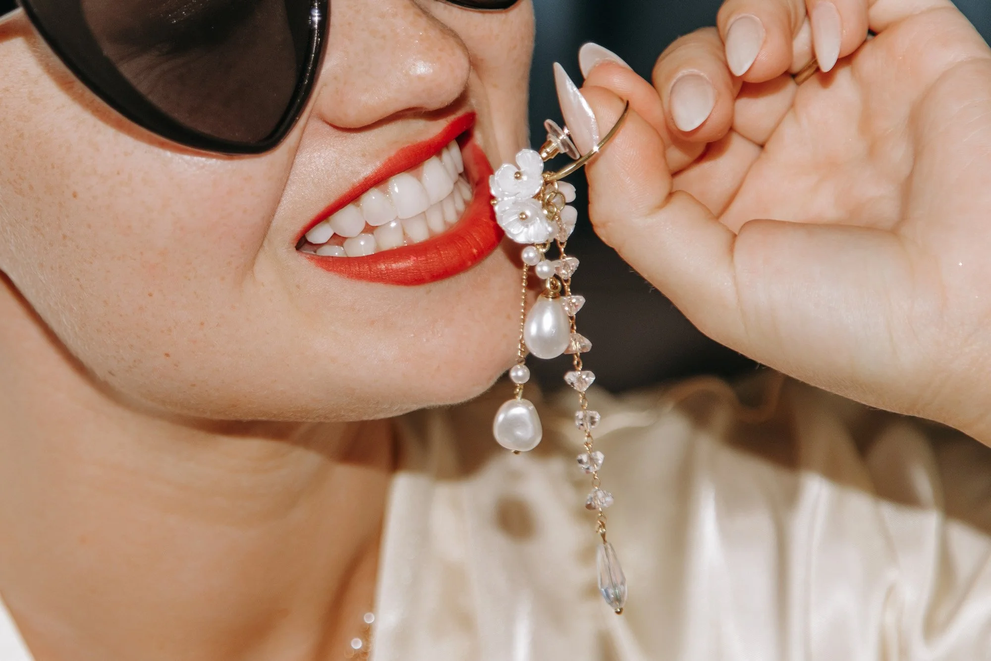 A woman smiling with red lipstick, wearing sunglasses, holding a pearl and floral-shaped earring near her face.
