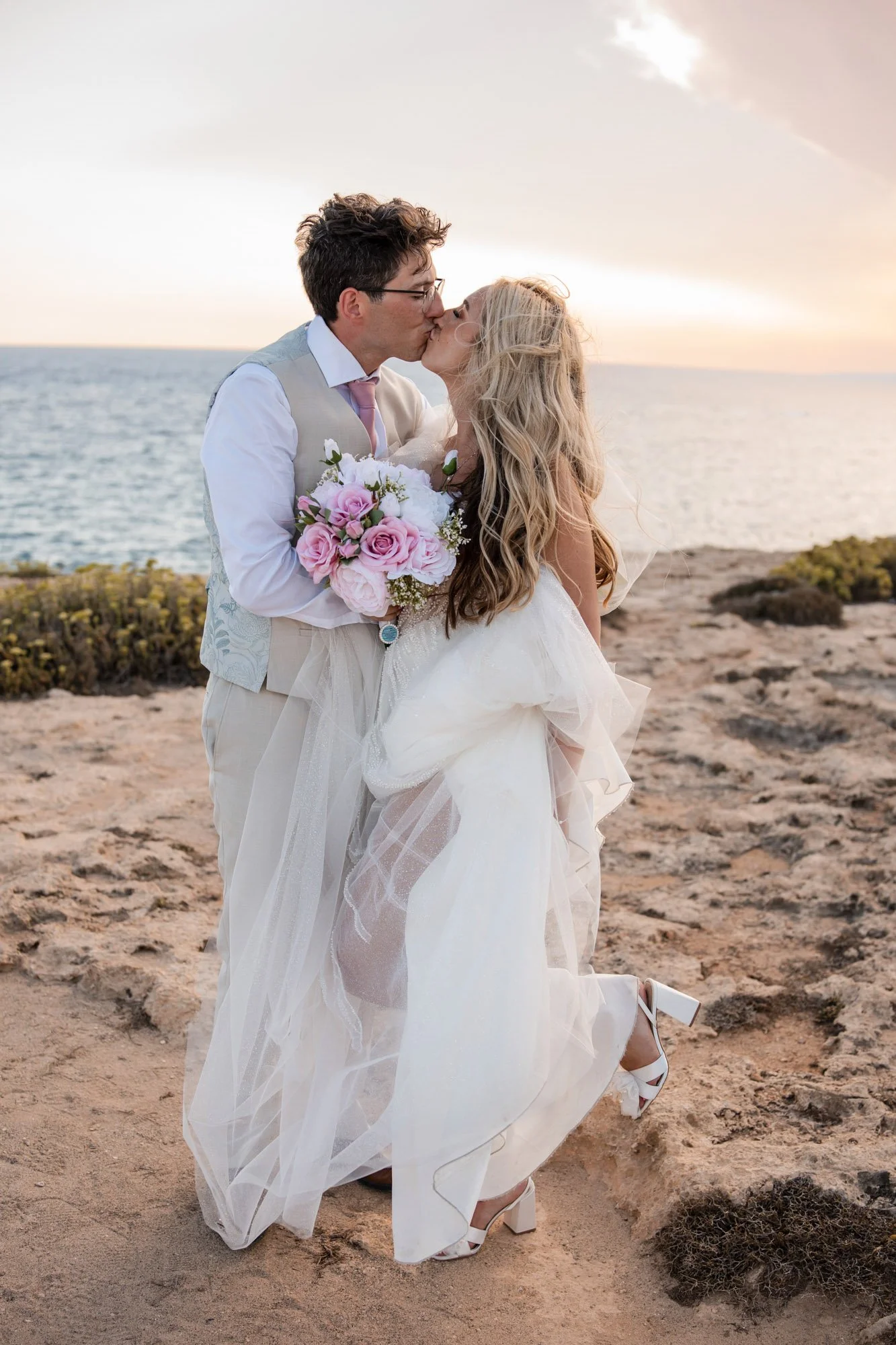 A couple dressed in wedding attire kissing on a rocky beach at sunset, with the ocean in the background.