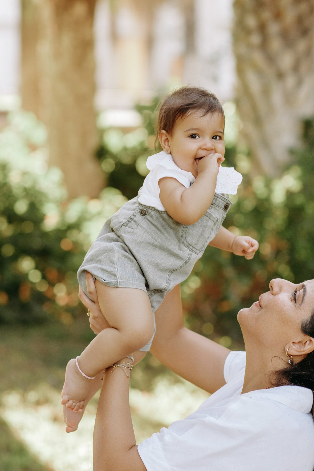 A woman lifting a young girl in a park with trees in the background, both smiling and looking happy.