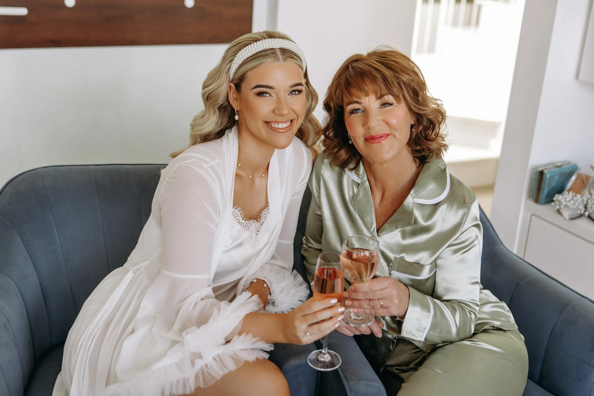 A young woman and older woman sitting on a dark gray sofa, smiling and holding glasses of rosé wine, celebrating together in a bright room.