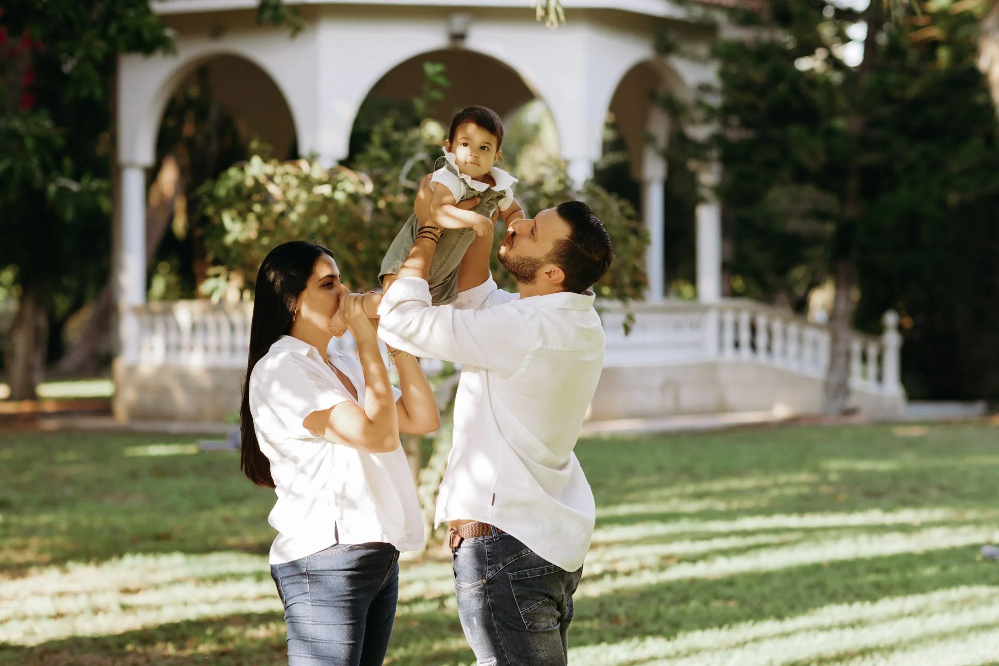 A family outdoors in front of a white gazebo with arches, with a man lifting a toddler girl, and a woman kissing the child's hand, all smiling.