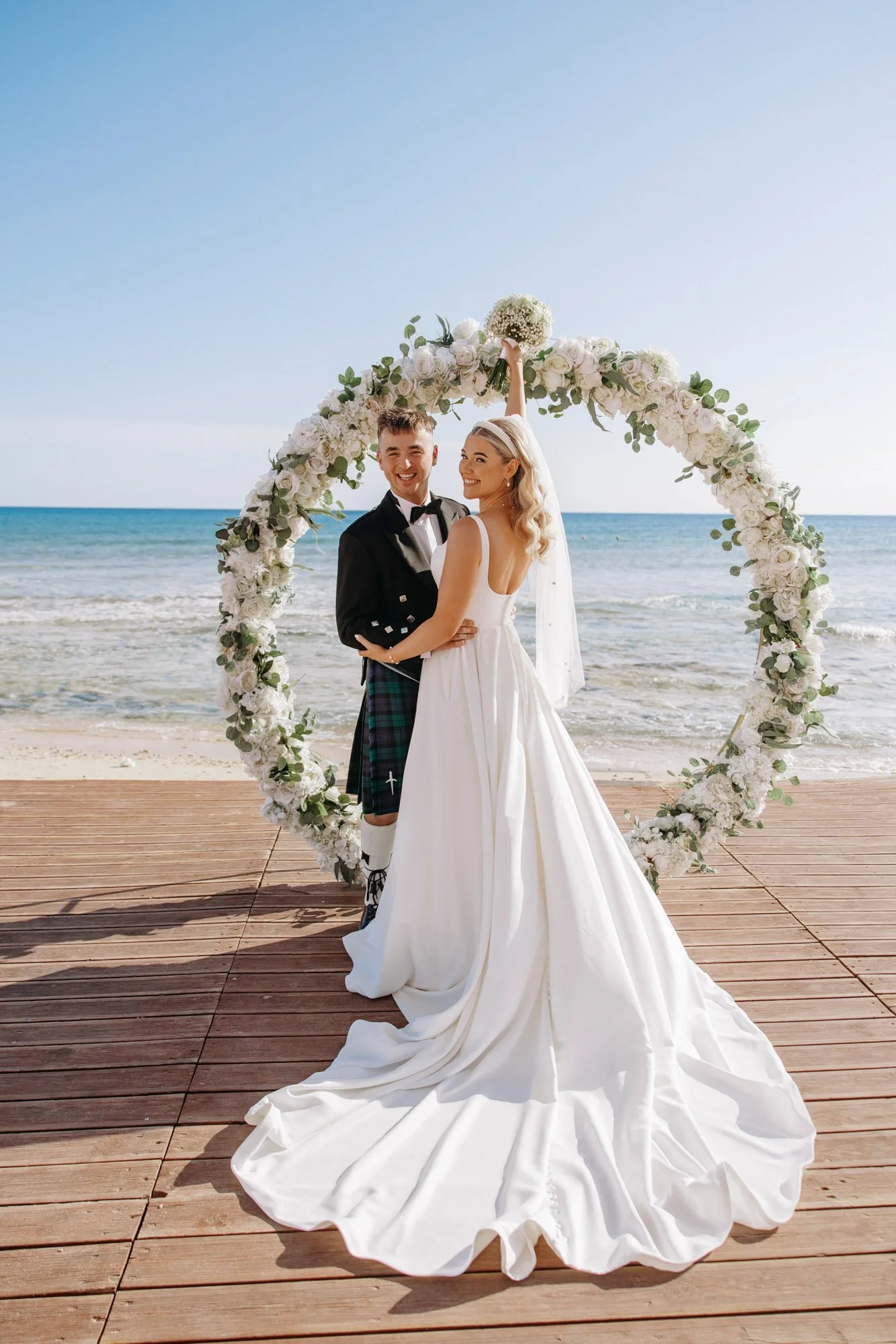 A couple in wedding attire standing on a wooden deck by the beach with a large heart-shaped floral arch behind them.