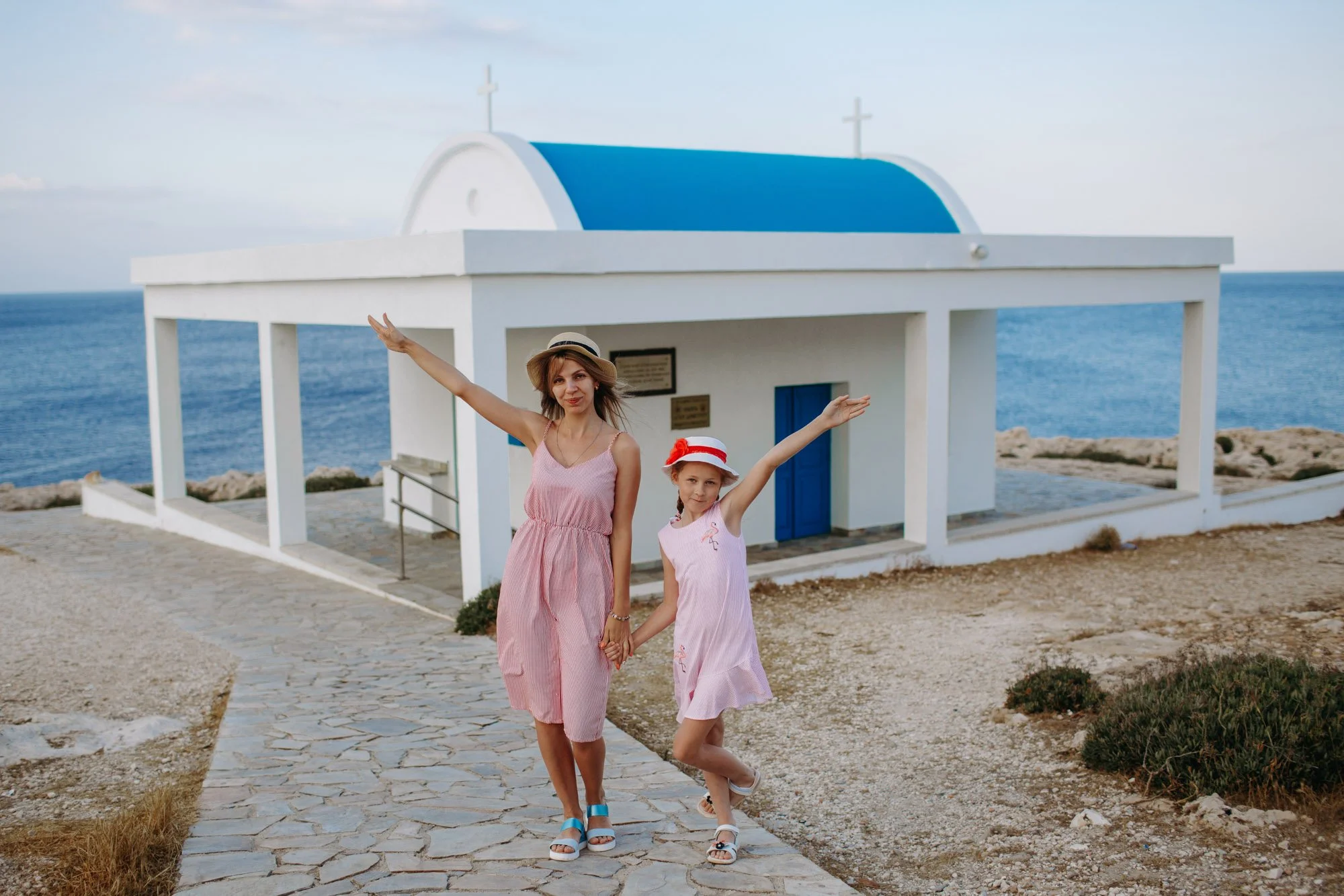 A woman and a girl, both dressed in pink dresses with hats, are holding hands and standing on a cobblestone pathway near the sea, with a small white chapel with a blue roof behind them.