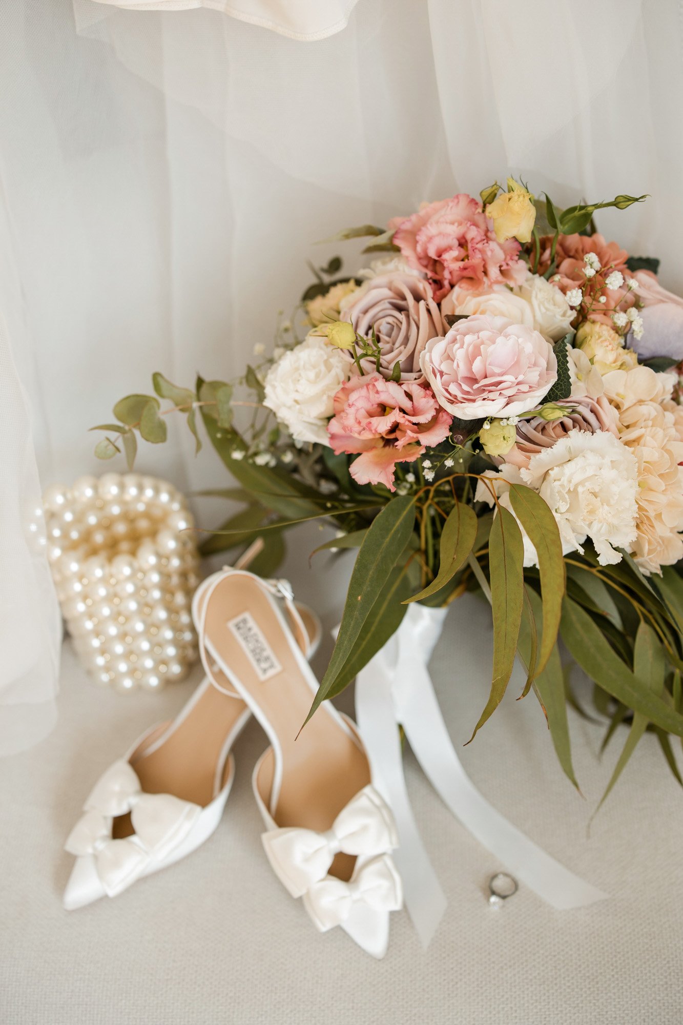 A wedding bouquet of pink, white, and lavender roses with greenery, a pair of white high-heeled shoes with bows and straps, and a pearl clutch purse with a diamond ring beside it.