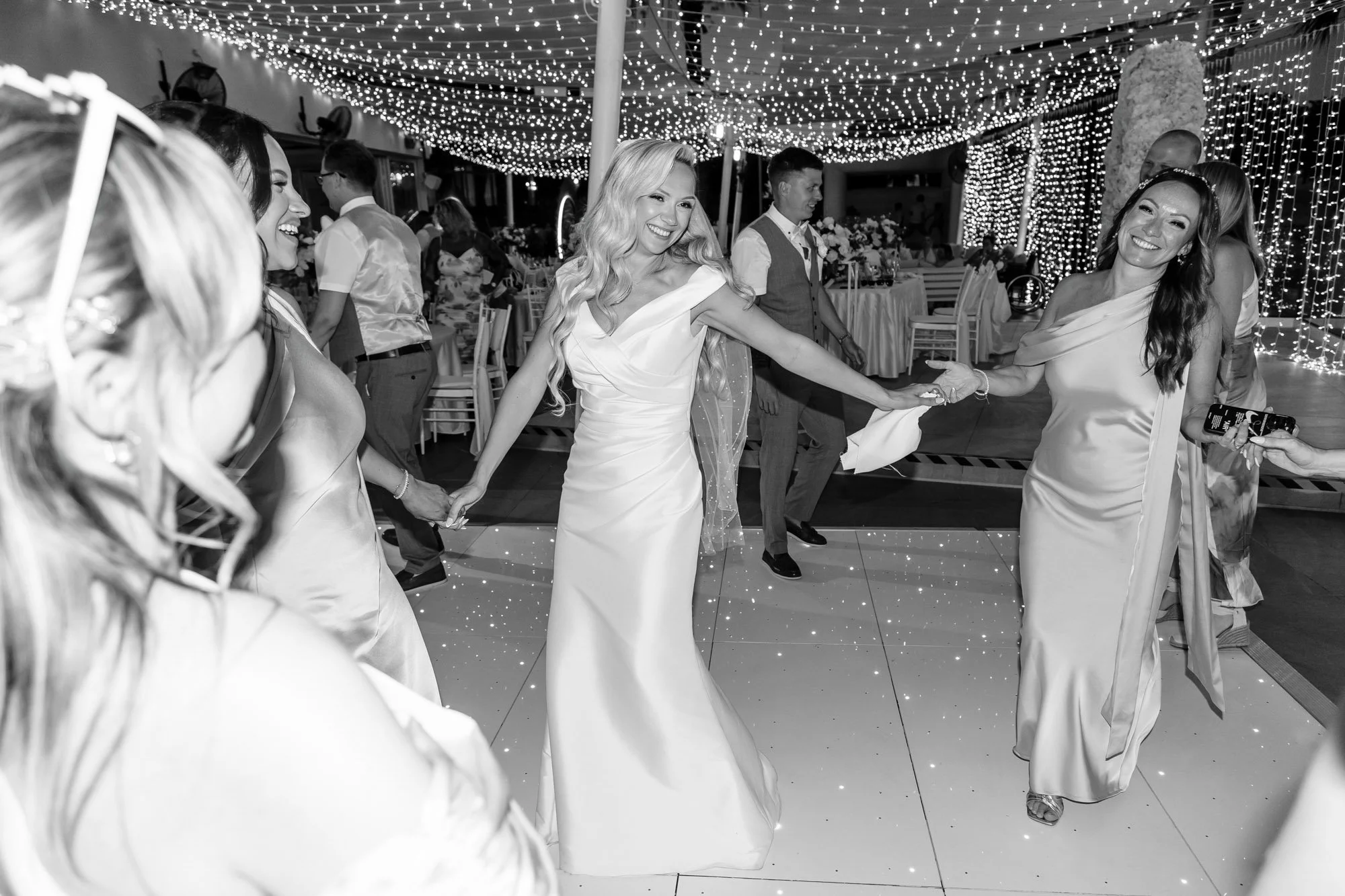 Women in elegant dresses dancing and holding hands at a wedding reception decorated with string lights and string curtains.