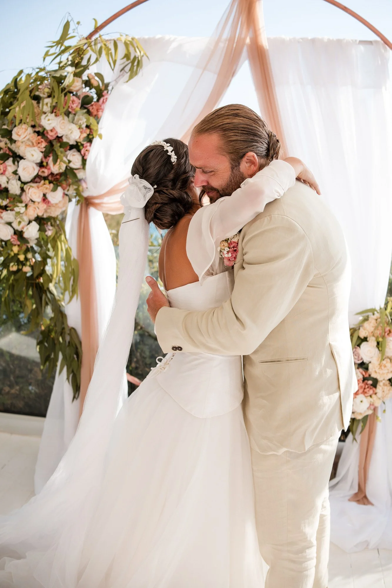 A bride and groom embrace during their wedding ceremony under a floral arch with pink and white flowers.