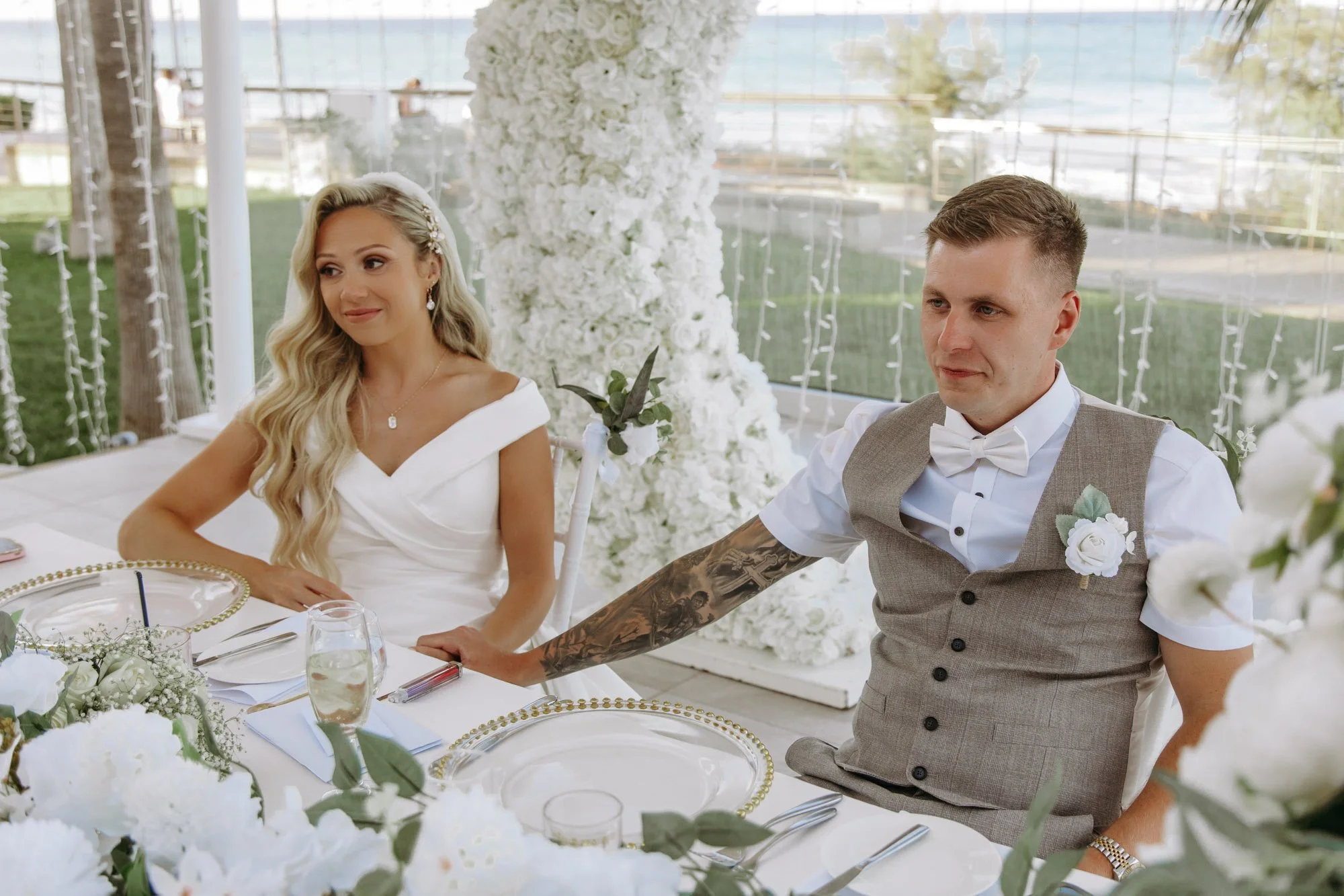 Bride and groom sitting at a wedding reception table decorated with white flowers and greenery, with the bride wearing a white dress and the groom in a grey suit vest with a white shirt and bow tie, outdoors with a large floral arrangement in the bac