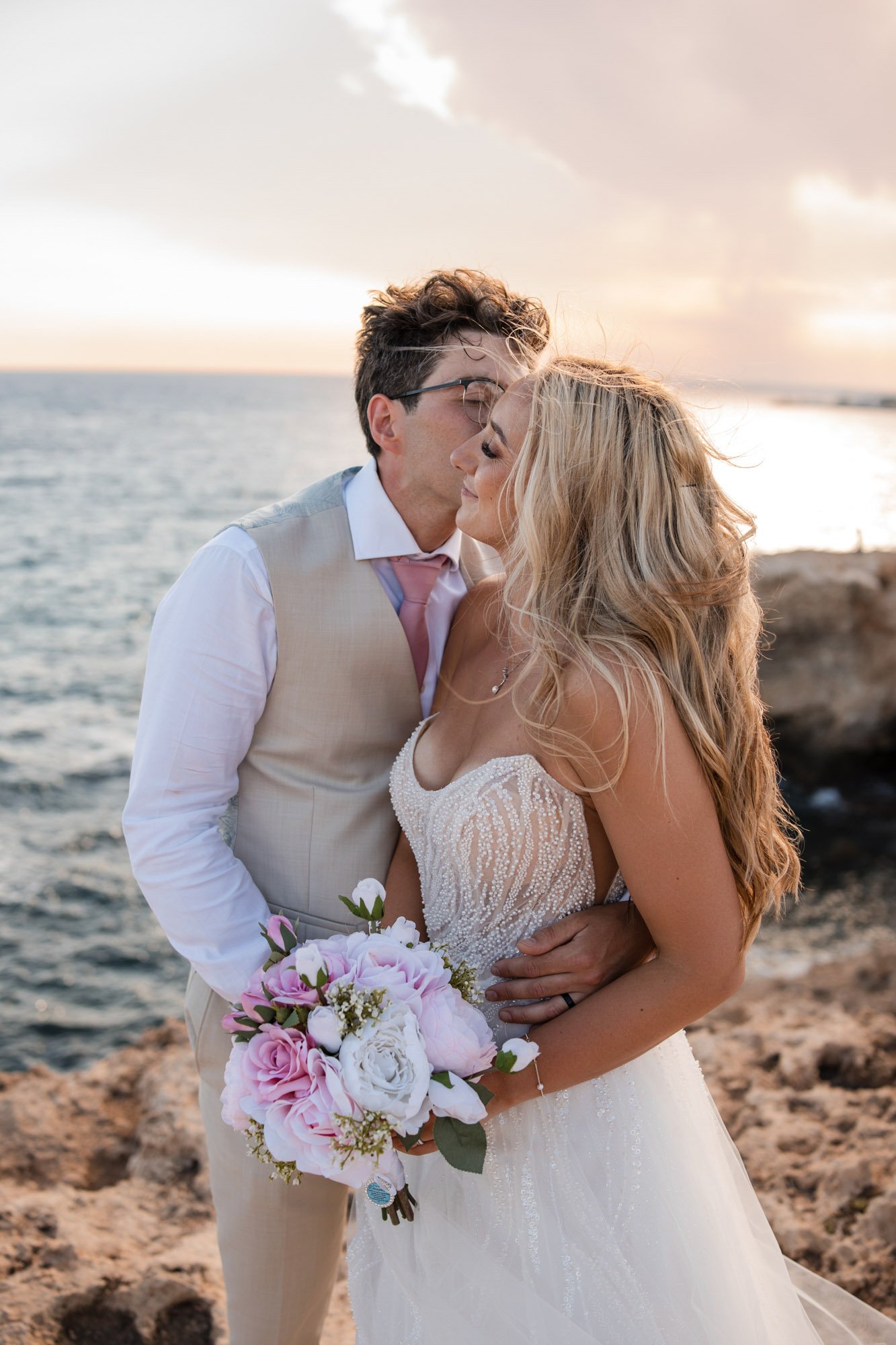 A newlywed couple sharing a kiss by the ocean at sunset, the bride holding a bouquet of pink and white flowers.