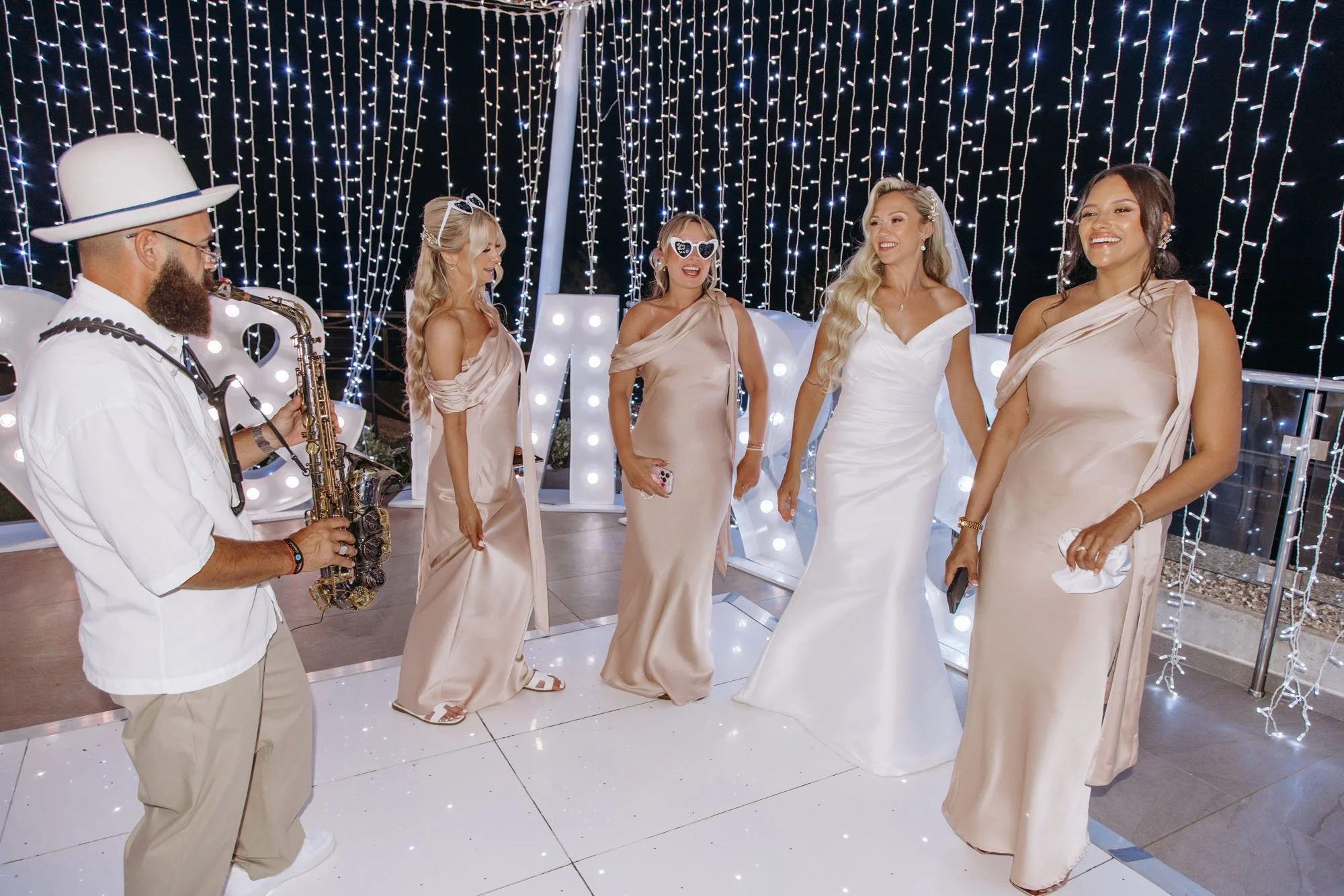 A group of five women in elegant wedding dresses standing together at a wedding celebration, with a man playing a saxophone. The background is decorated with string lights and large illuminated letters.