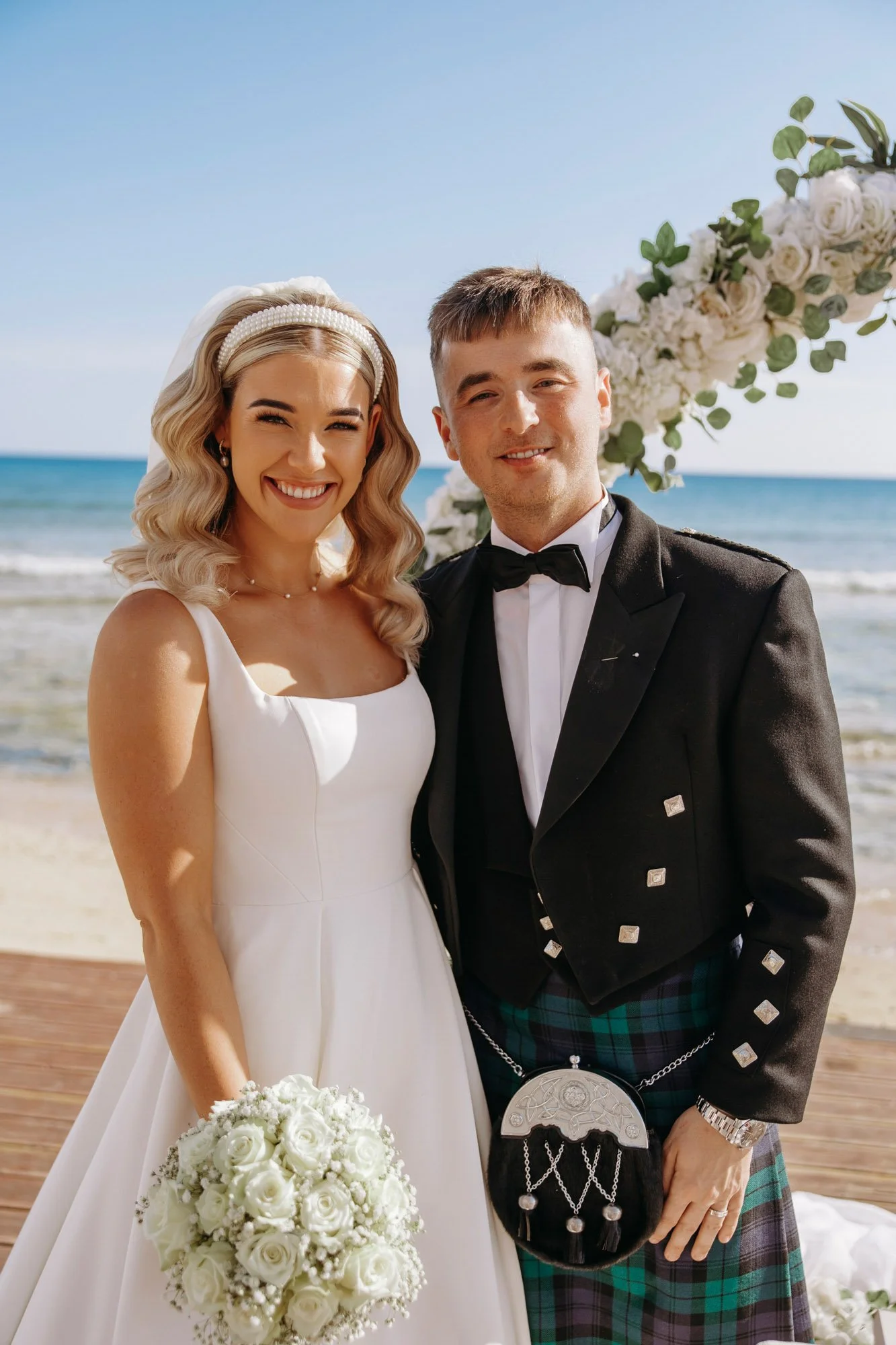 A bride and groom posing together on a beach during their wedding, smiling, with the ocean and sky in the background. The bride wears a white wedding dress and holds a bouquet of white roses, while the groom wears formal attire with a kilt and black 